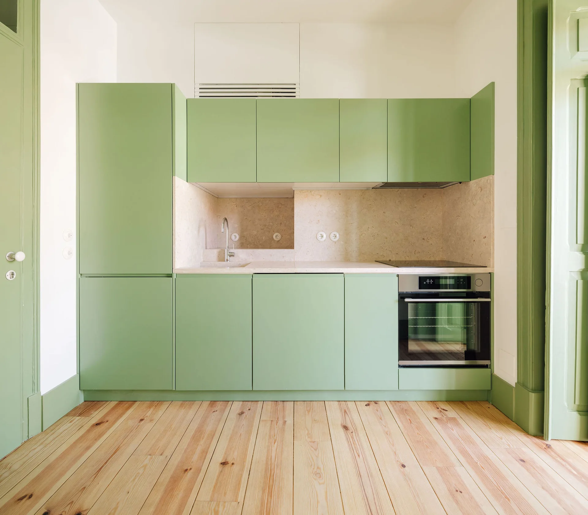 Minimalist kitchen featuring matte sage green cabinetry, a stone-textured backsplash, built-in appliances, and light wood plank floors.
