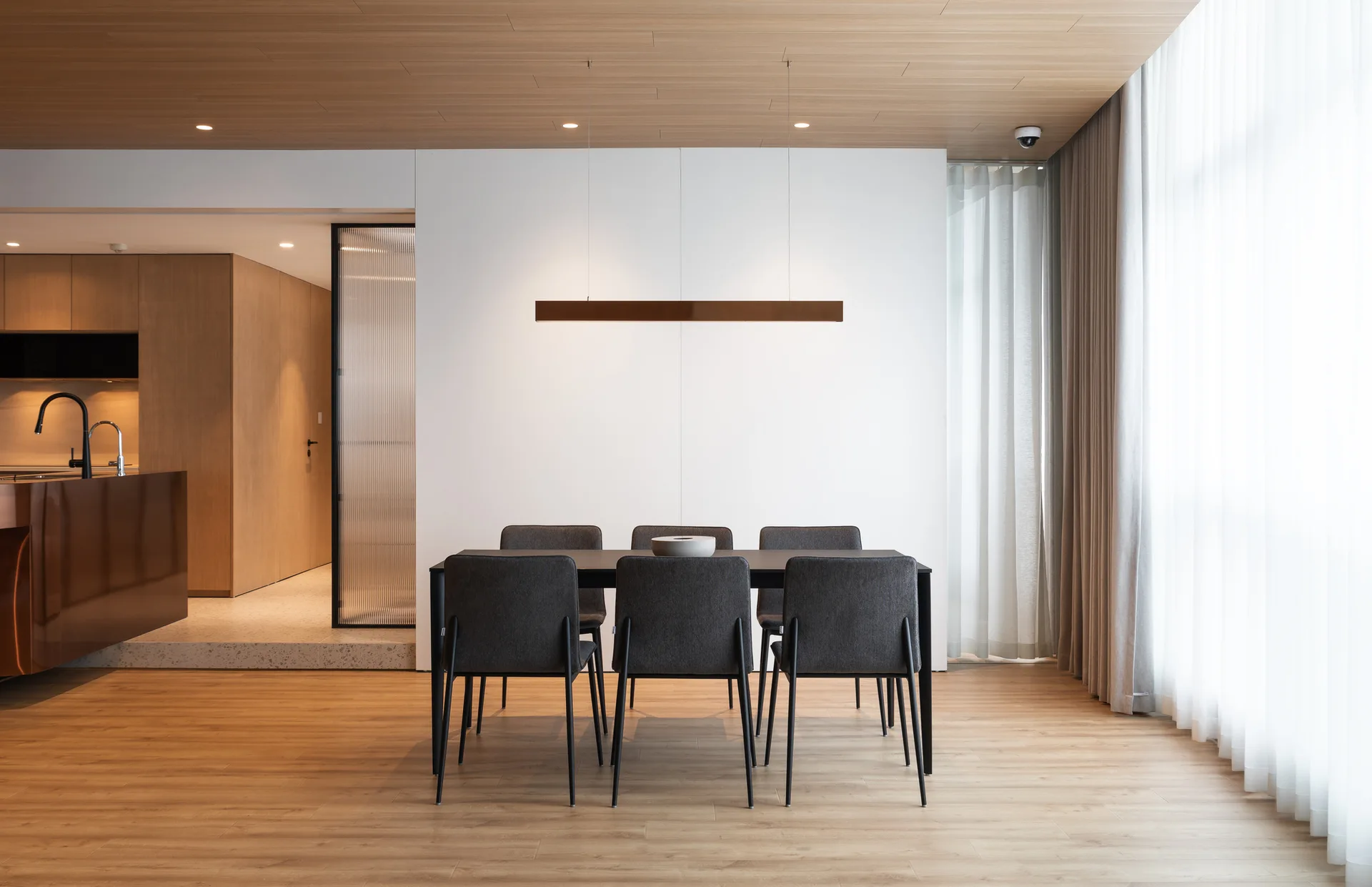 A modern dining room features a black table with charcoal chairs under a wood-paneled ceiling and a linear pendant light.