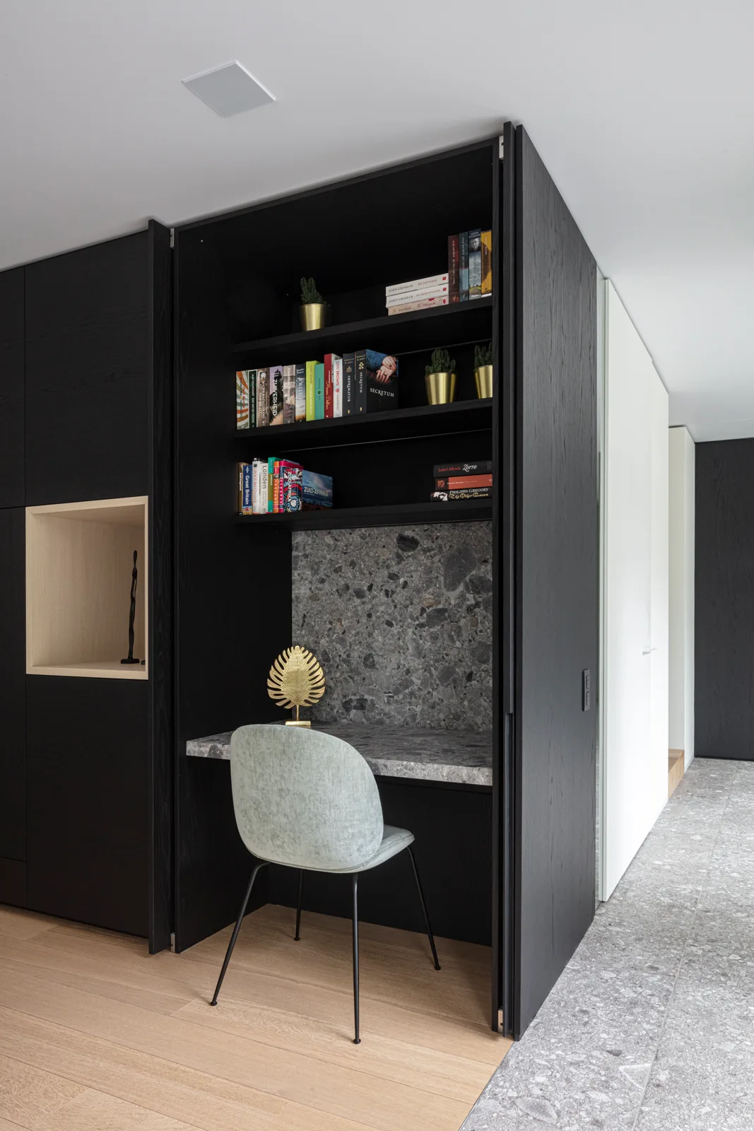 A modern home office nook built into black wood cabinetry features a gray stone desk, built-in shelving with books, and a light gray chair.