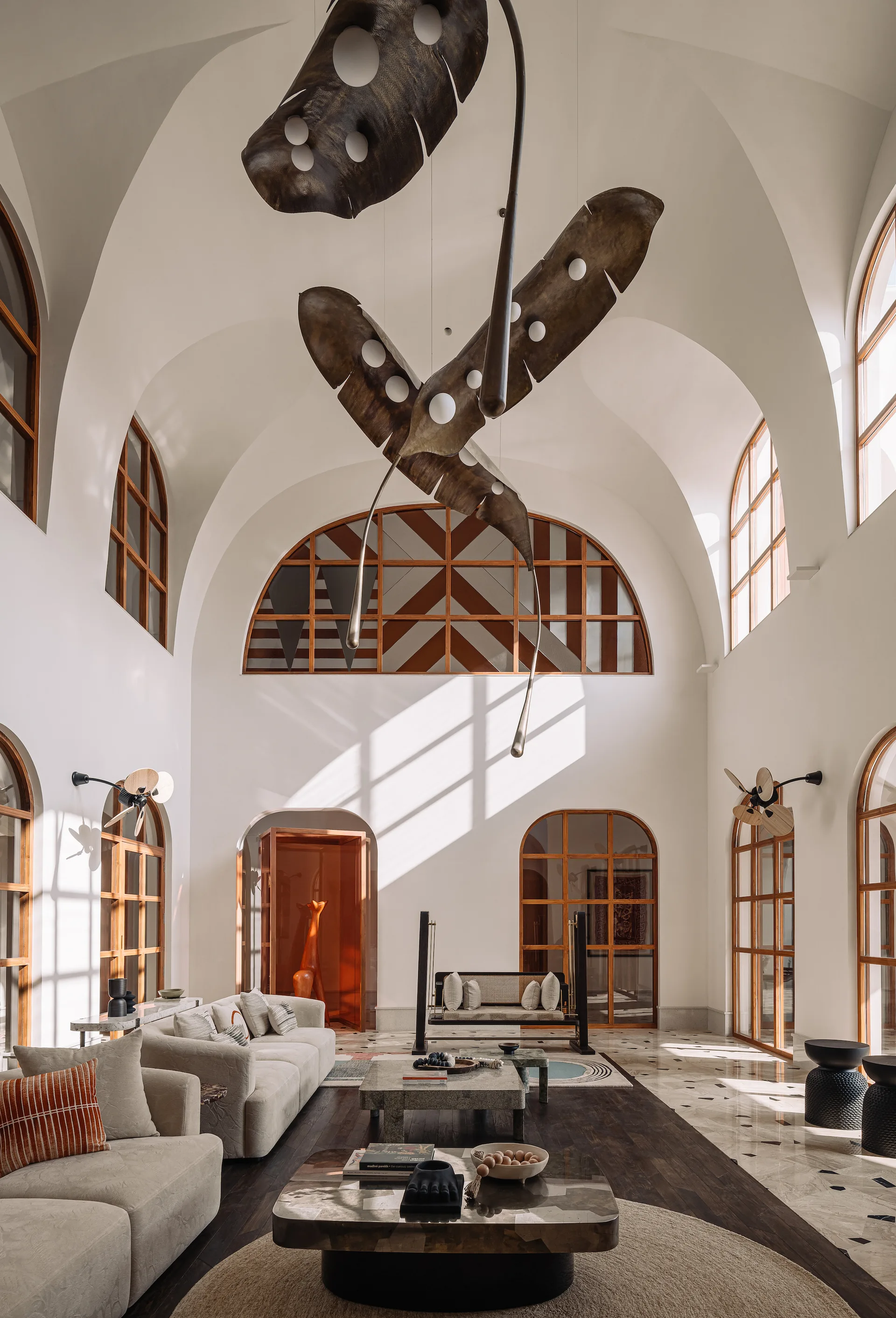 A spacious living room features high vaulted ceilings, an oversized leaf-shaped pendant light, cream sofas, and large arched wooden windows.