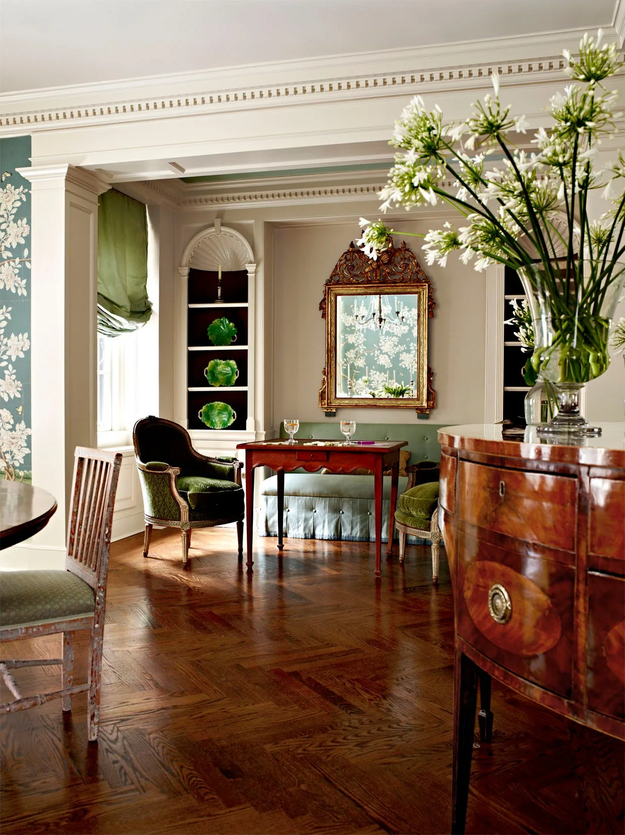 A traditional dining room featuring polished herringbone wood floors, a small wooden table with green velvet chairs, and an arched built-in shelving alcove.