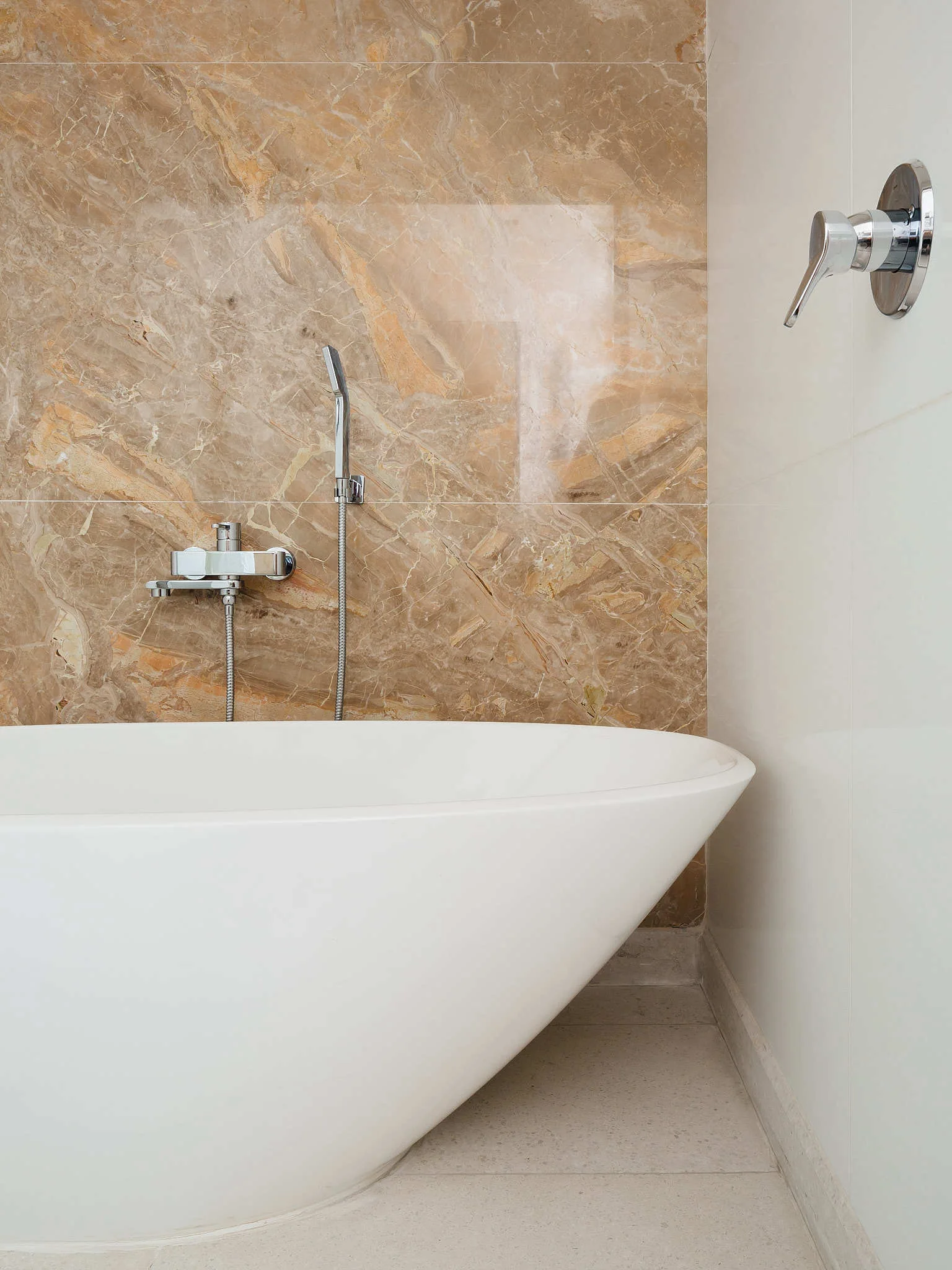 A white freestanding bathtub sits against a polished brown marble accent wall and white panels with chrome bathroom fixtures.