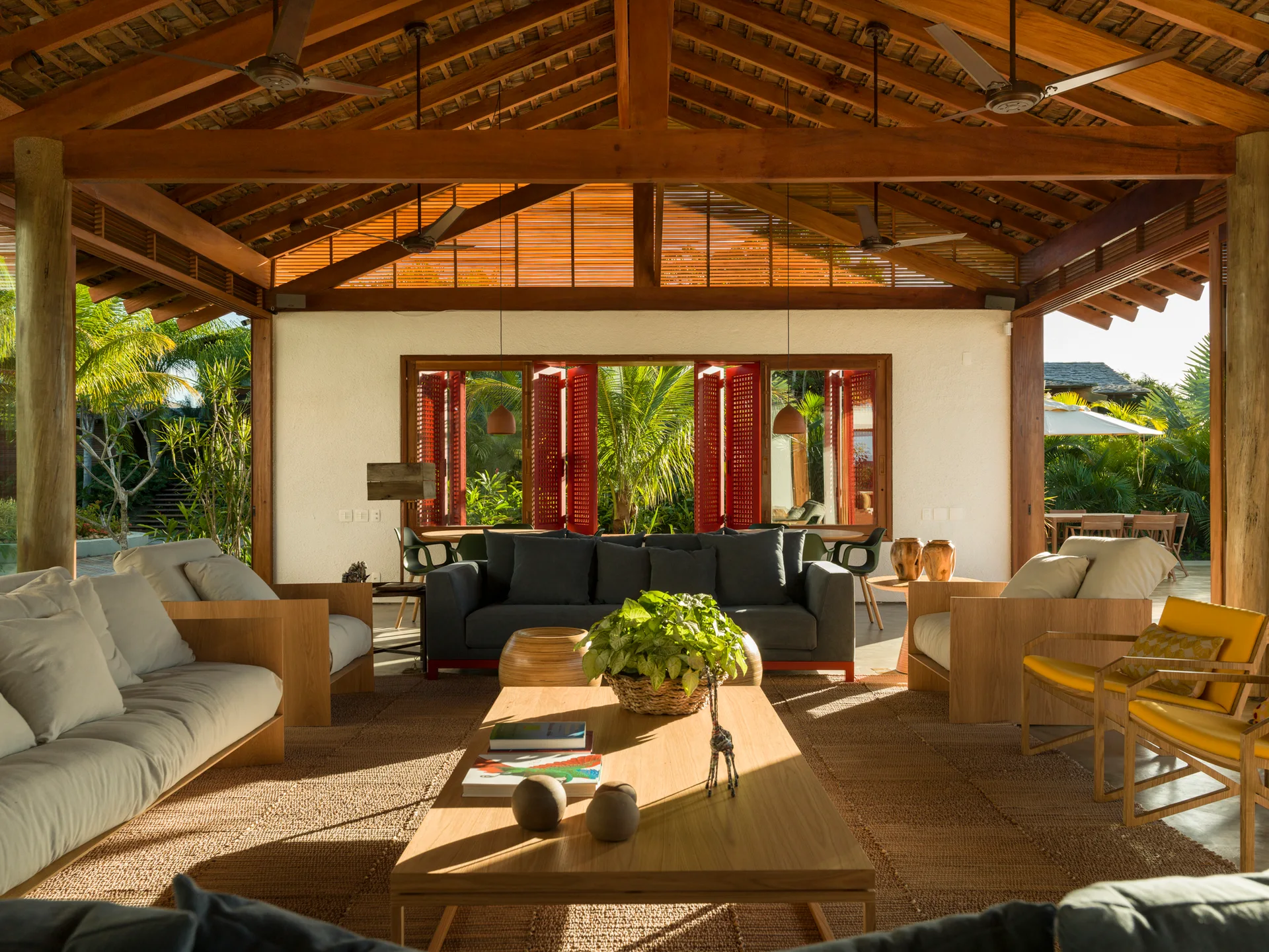 An open-air living room with a vaulted wood beam ceiling, neutral-toned sofas, a wooden coffee table, and large window openings framing tropical greenery.