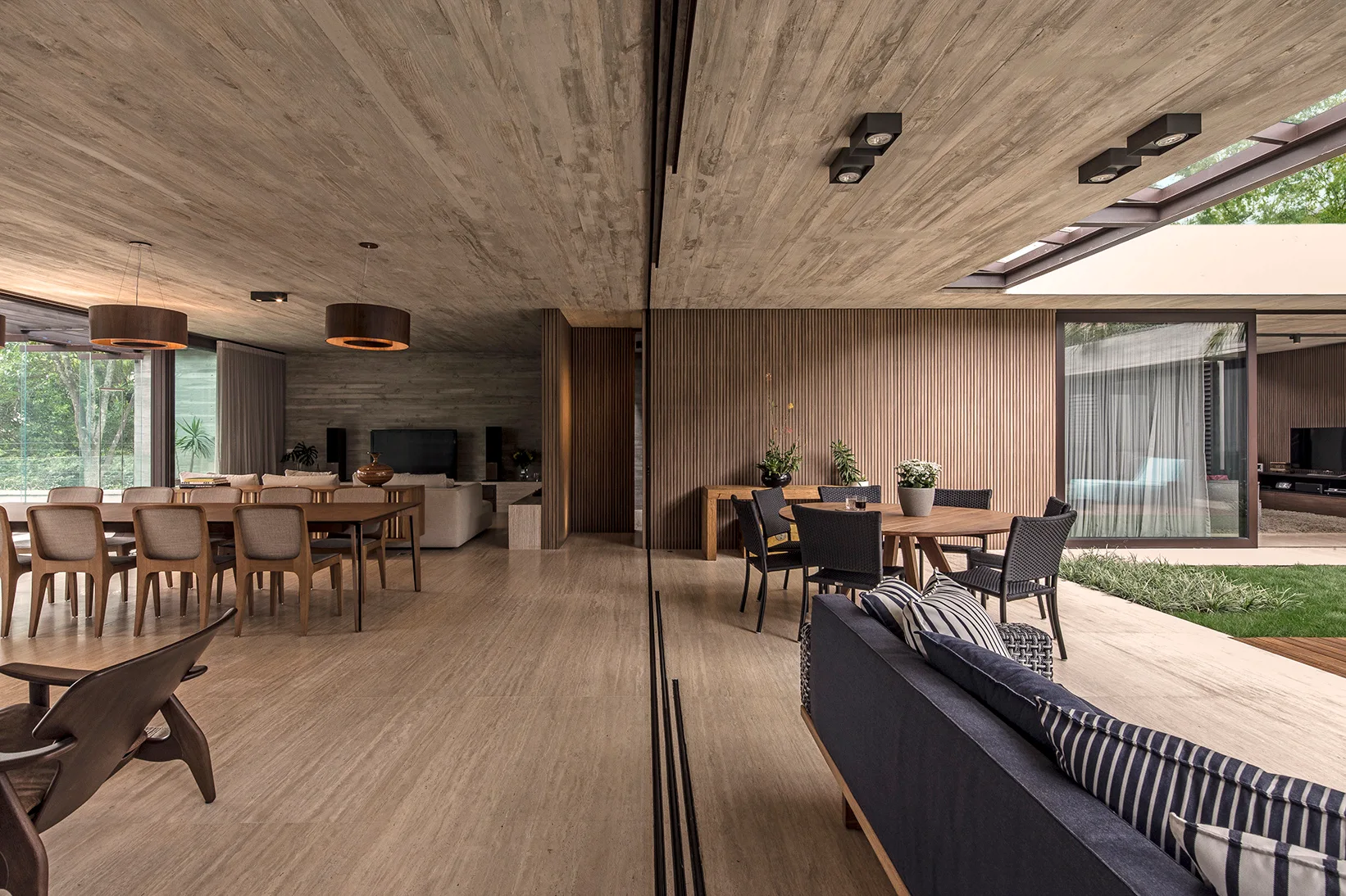 A modern open-plan living and dining area features a board-formed concrete ceiling, vertical wood slat wall paneling, and large sliding glass doors.