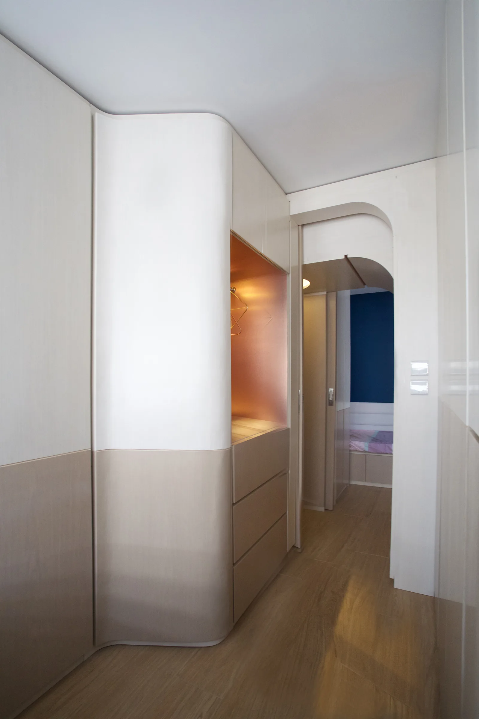 Minimalist hallway featuring custom beige and white cabinets with a curved corner, an illuminated open niche, and a matching arched doorway.