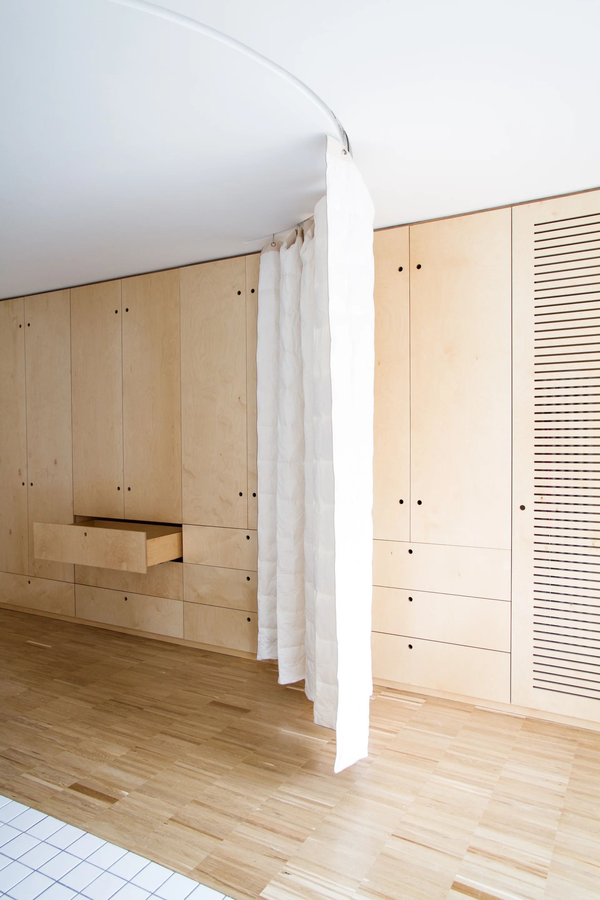A light wood built-in wardrobe features integrated circular handles and a white partition curtain hanging from a curved ceiling track.