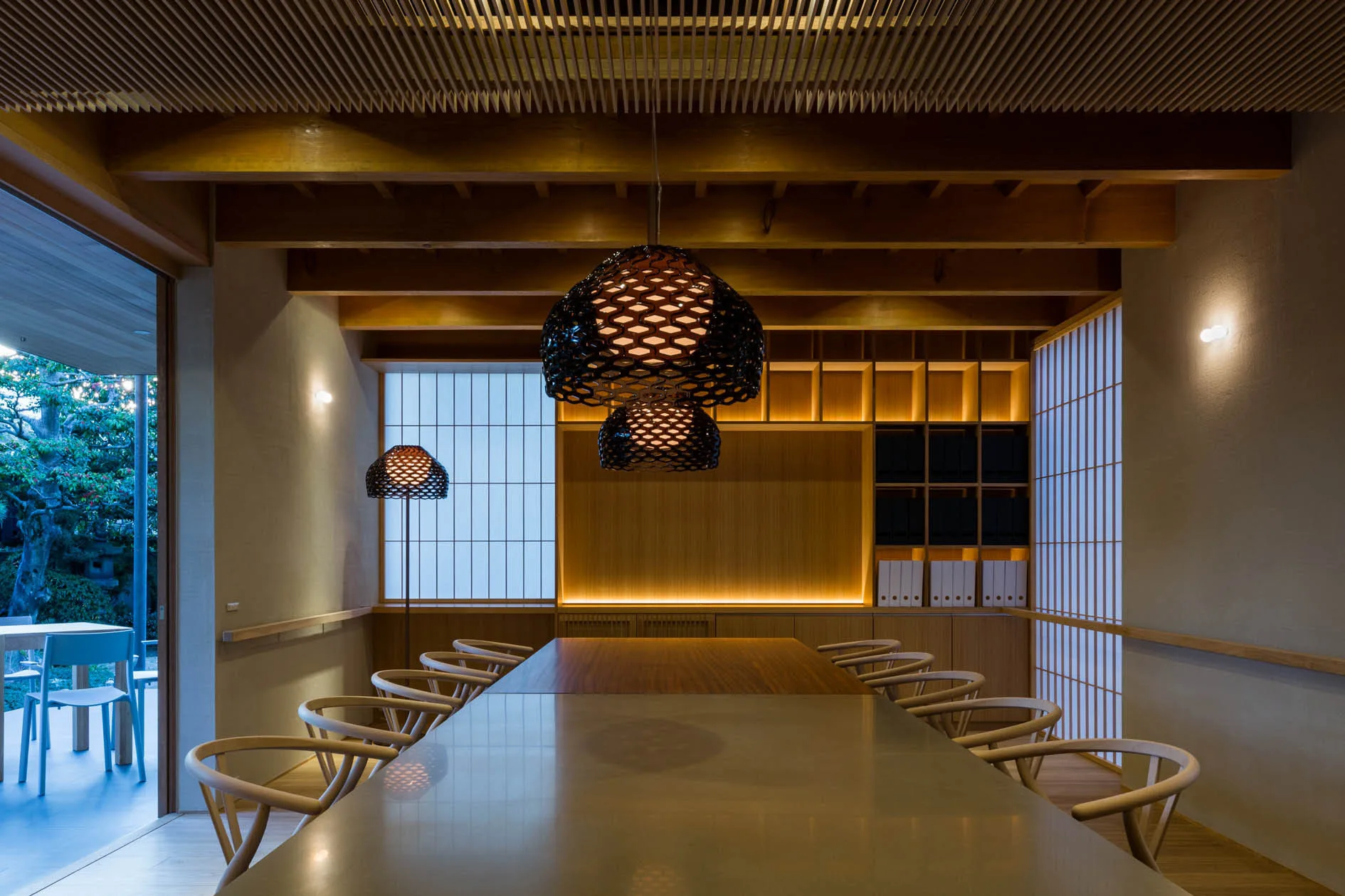 A long wooden dining table is surrounded by curved chairs under a slatted ceiling, illuminated by tiered black honeycomb pendant lights and shoji screens.