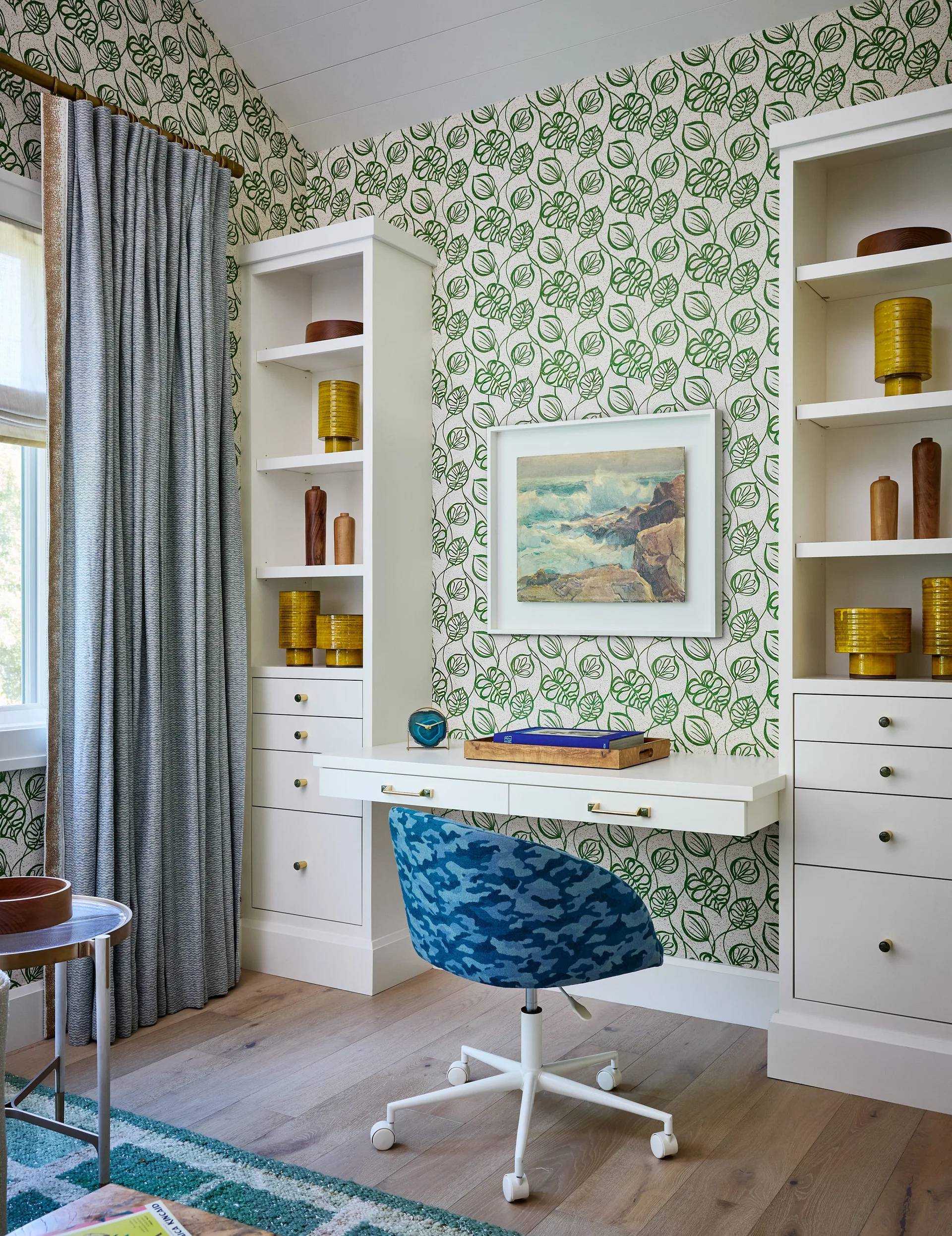 A home office features a white built-in desk and bookshelves against green leaf-patterned wallpaper with a blue patterned swivel chair.
