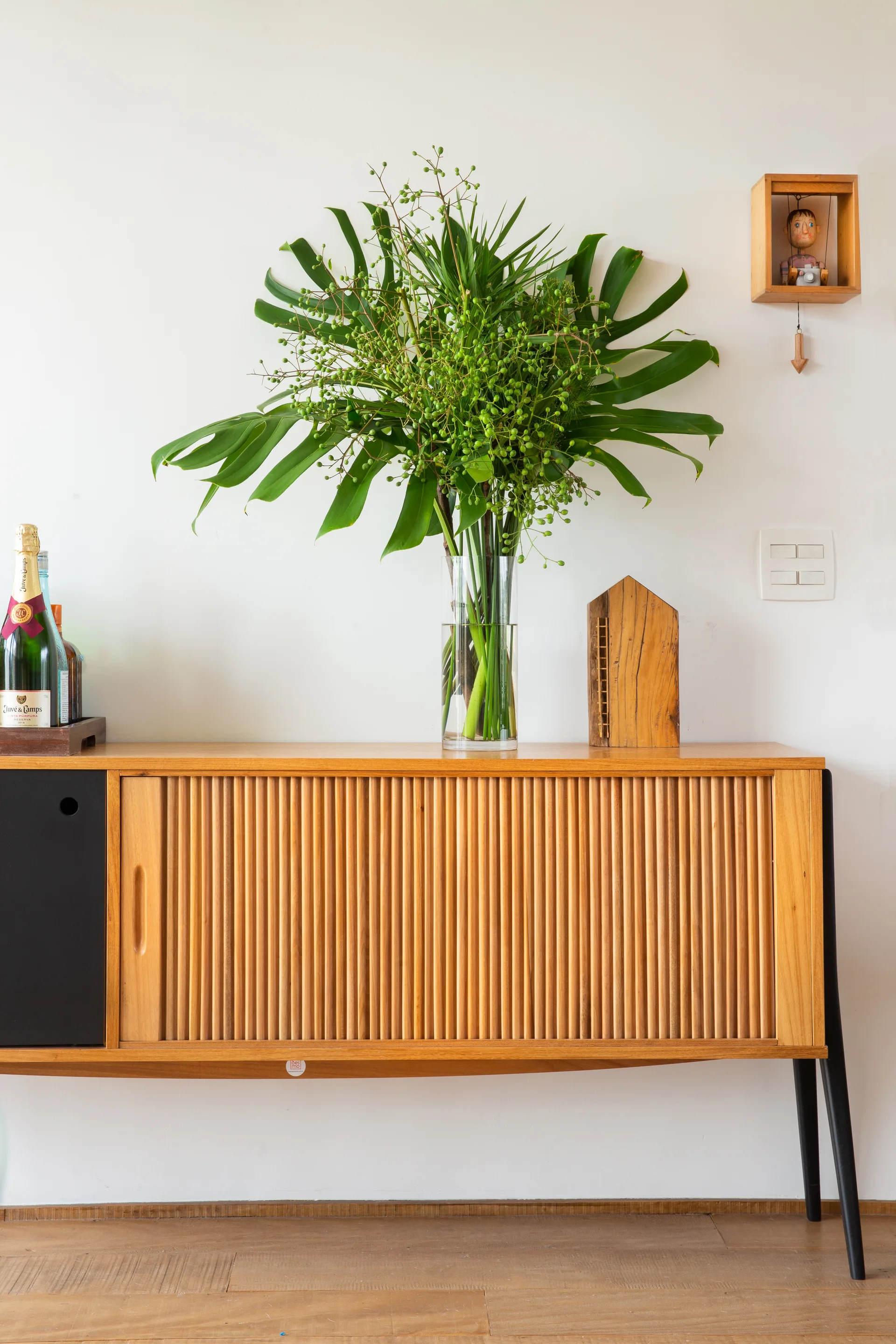A mid-century modern wooden sideboard with ribbed tambour doors features a clear glass vase holding large green leaves and berries against a white wall.