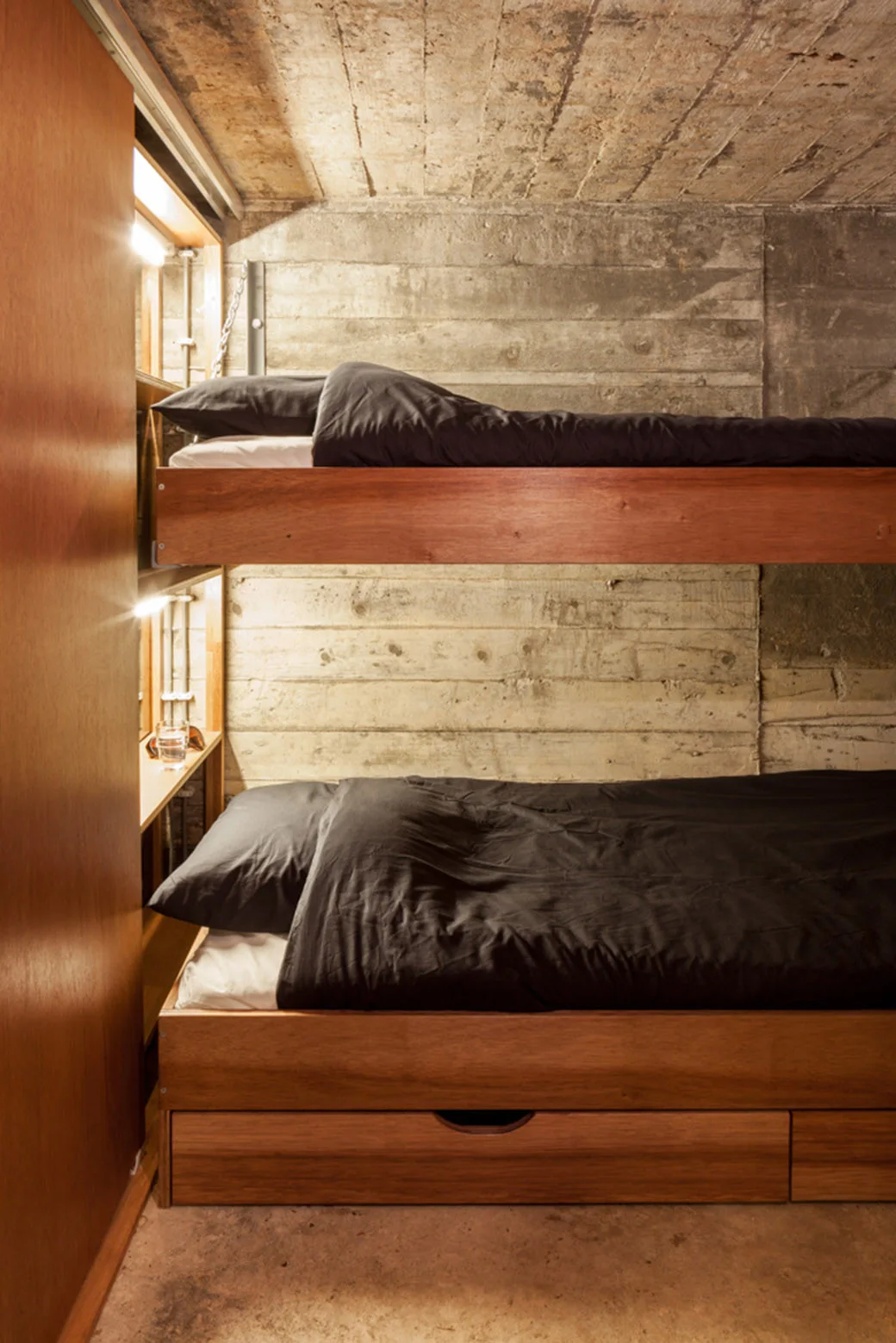 A wooden bunk bed with dark gray bedding sits against a board-formed concrete wall in a compact room with integrated warm lighting.