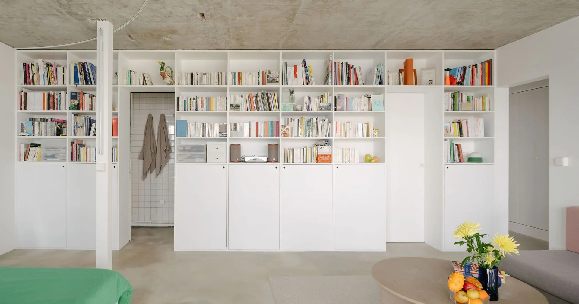 Large floor-to-ceiling white shelving unit filled with books is set beneath a raw concrete ceiling in a minimalist living room.