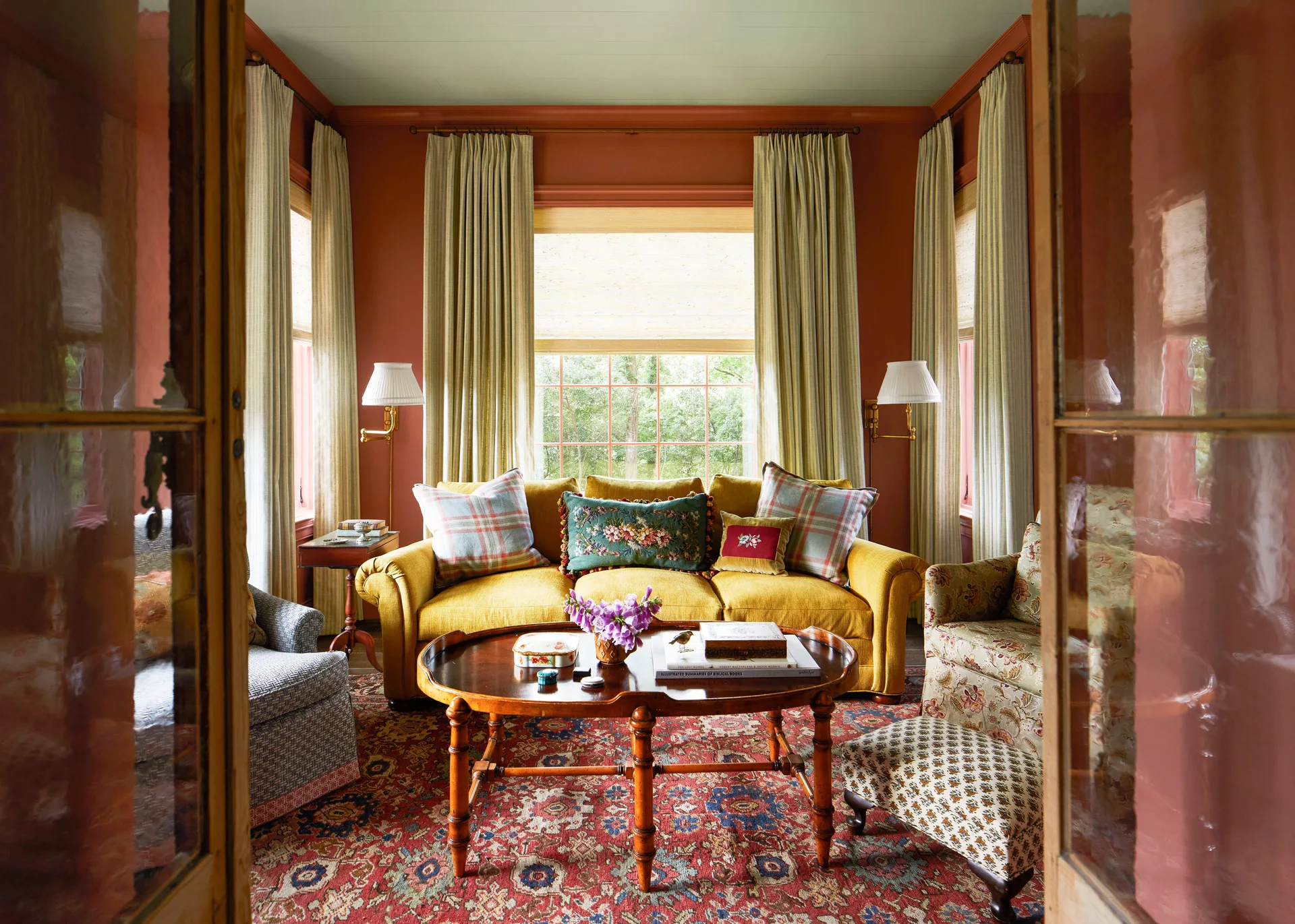 A traditional living room with a yellow sofa, an oval wood coffee table, and patterned armchairs against terracotta walls and a green ceiling.