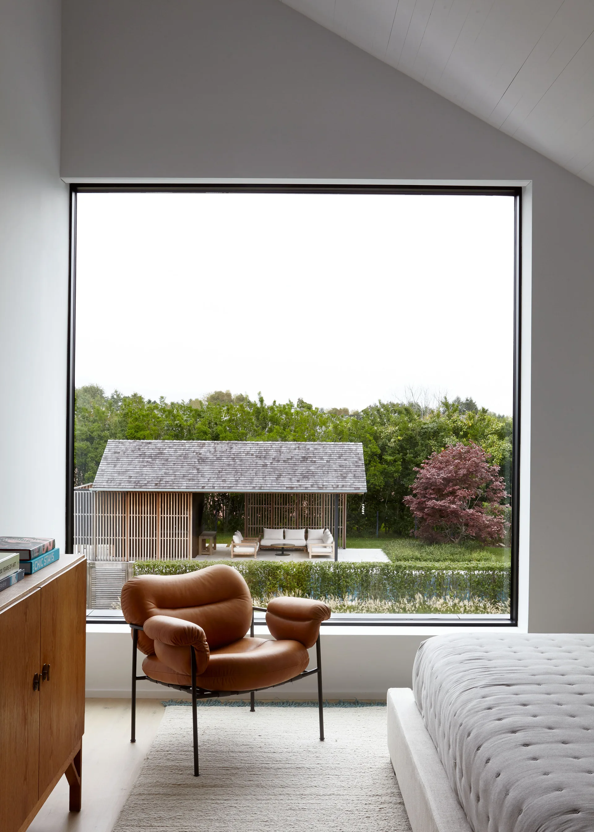 A modern bedroom features a large square window showing a garden pavilion, alongside a tan leather armchair and a gray tufted bed.