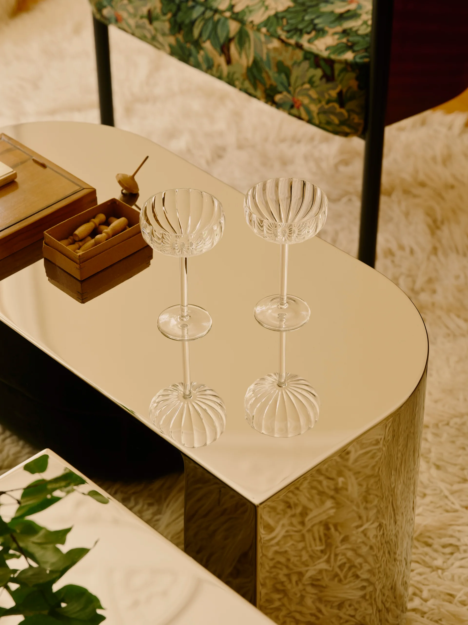 Two ribbed glass coupes sit on a polished mirrored coffee table, which rests on a high-pile shag rug next to an armchair.