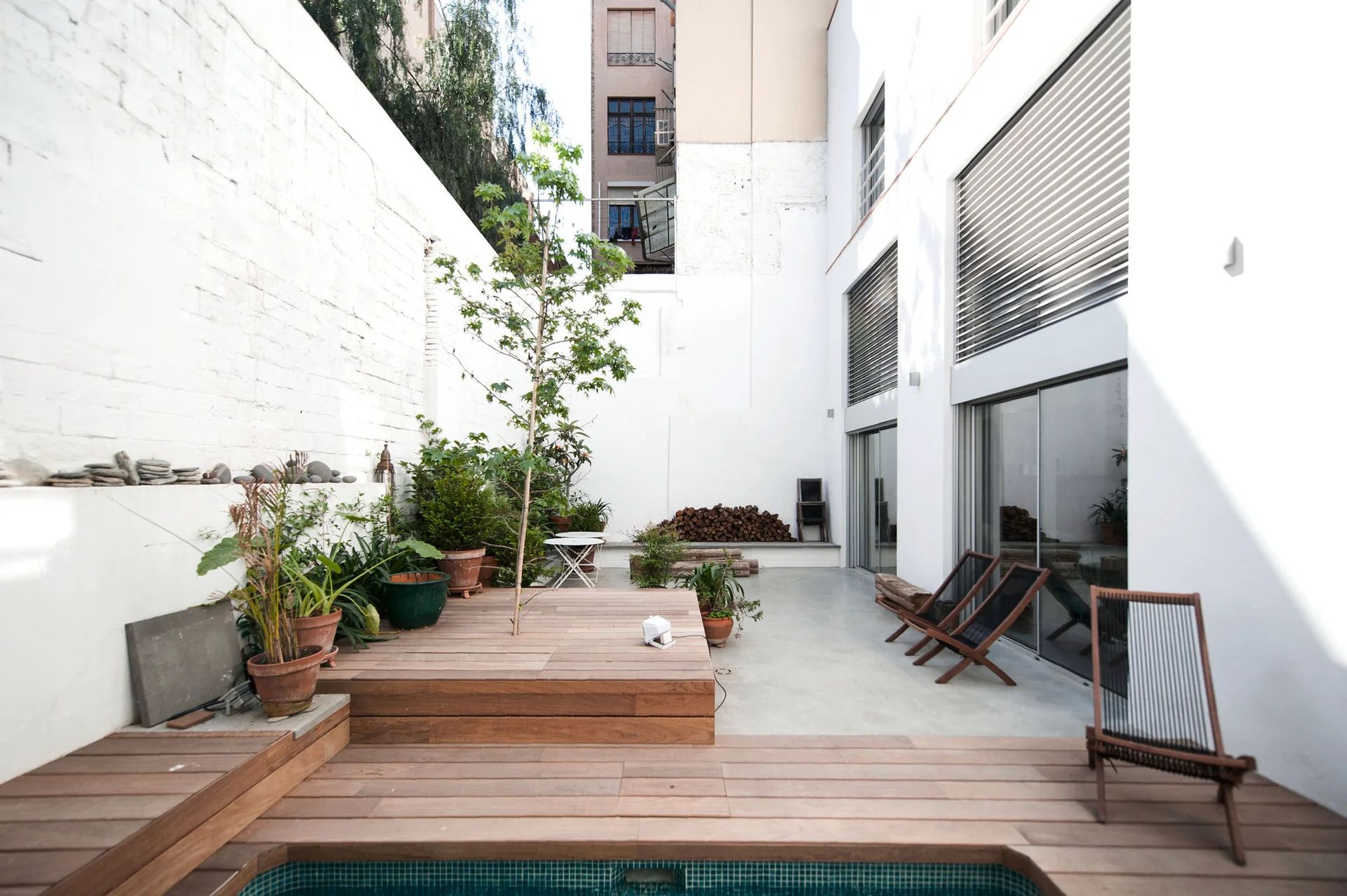 A minimalist outdoor courtyard featuring multi-level wooden decking, a small tiled pool, tall white walls, and potted plants.