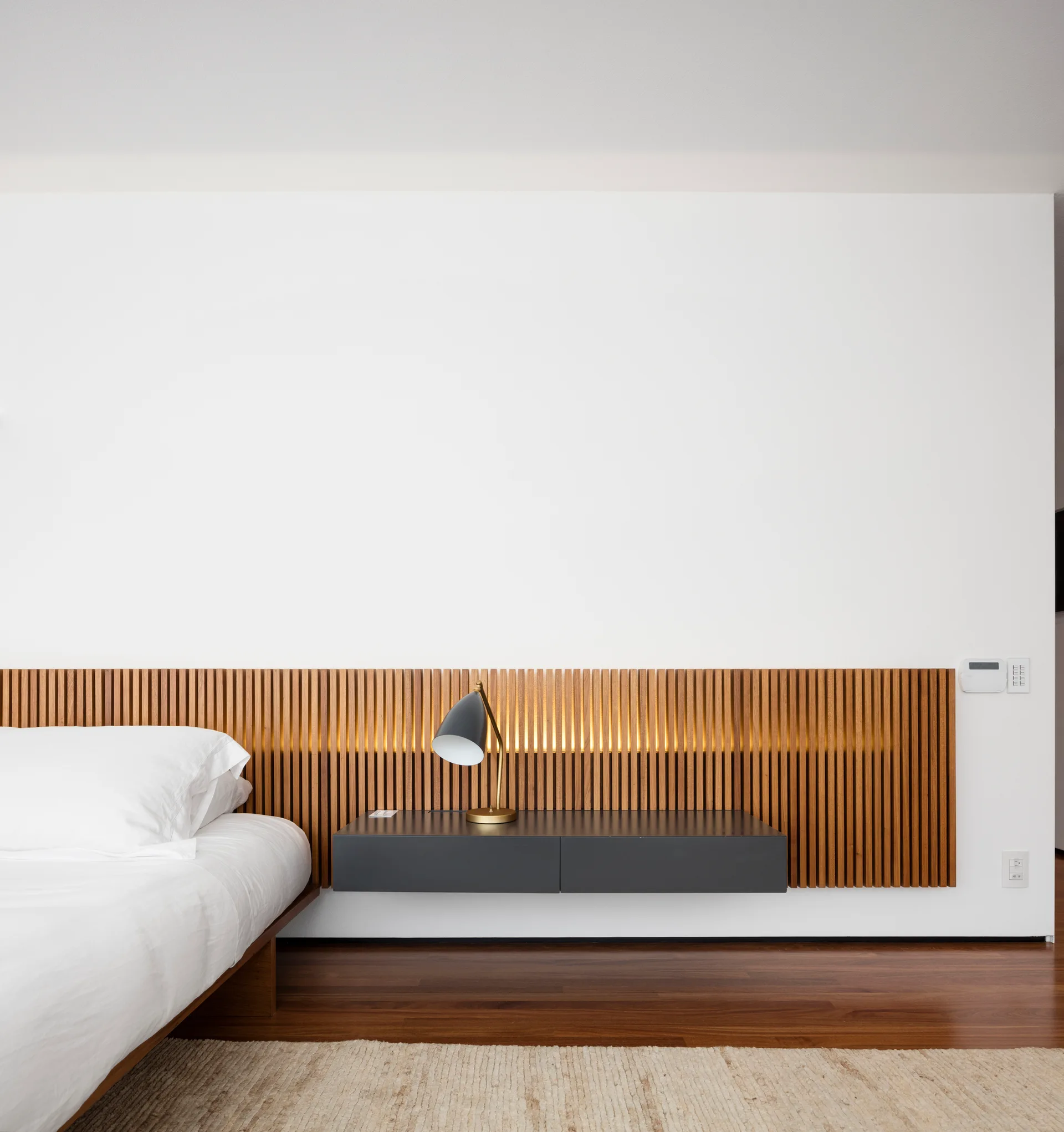 A minimalist bedroom features a floating charcoal nightstand beneath a backlit slatted wood headboard panel mounted on a plain white wall.