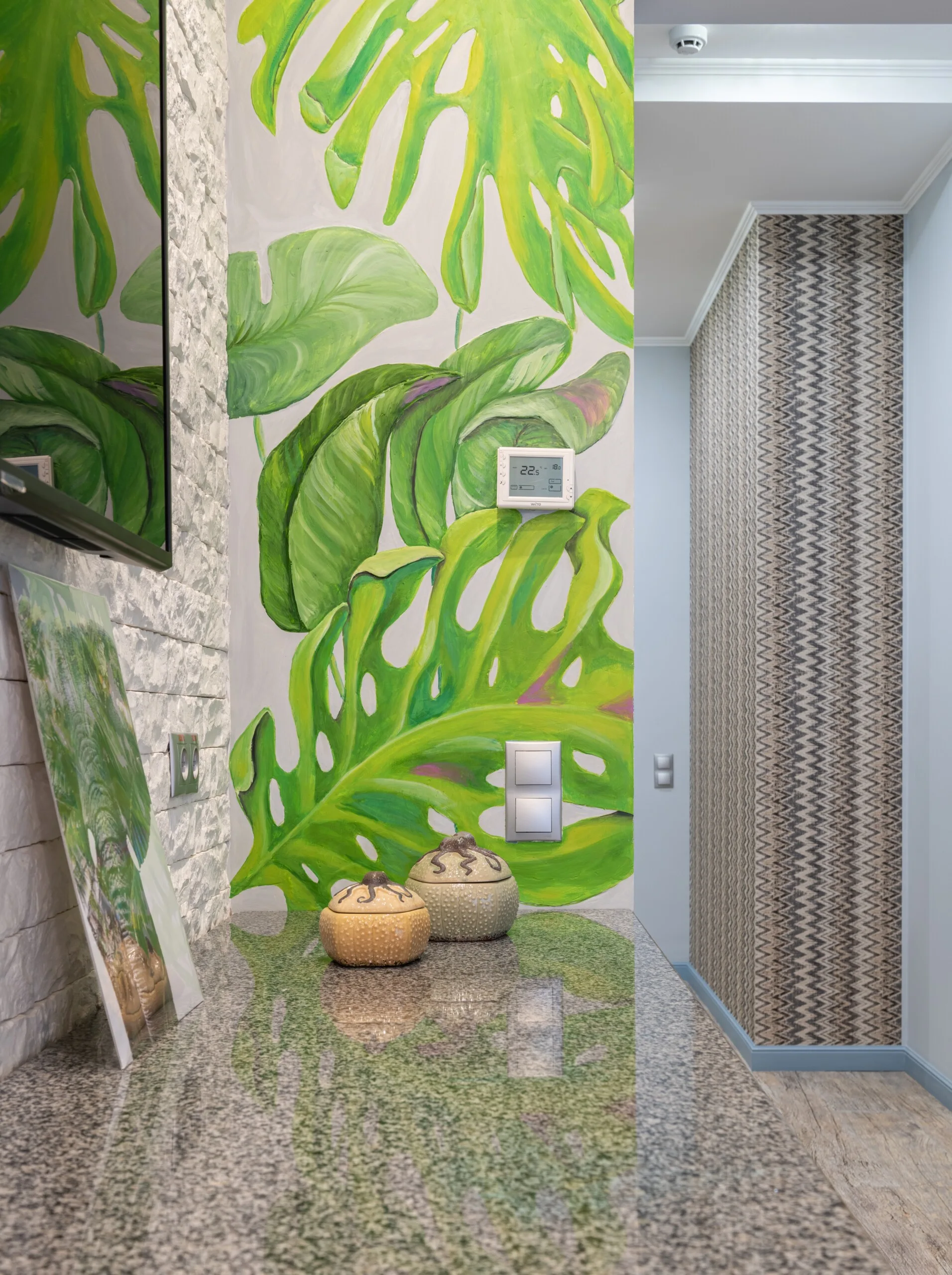A green monstera leaf wall mural stands next to a white brick-textured wall and a polished granite countertop with two decorative ceramic bowls.