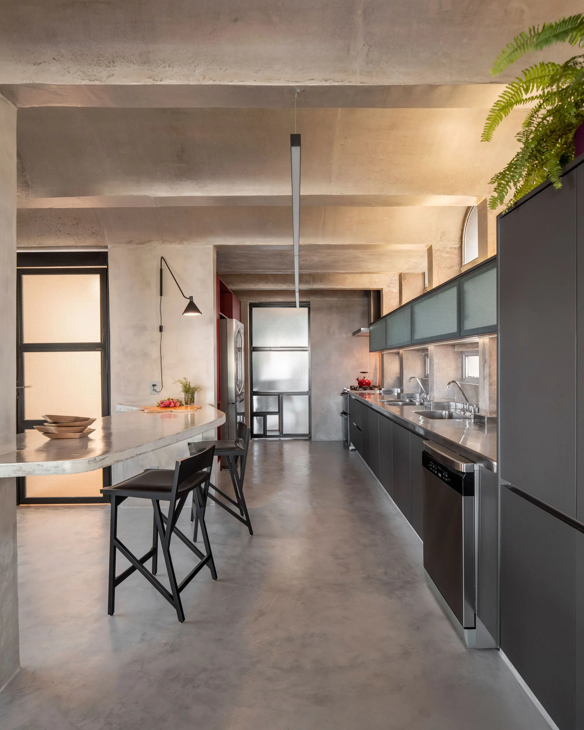 A modern industrial kitchen featuring an exposed concrete vaulted ceiling, a curved concrete island with black stools, and matte black cabinetry.