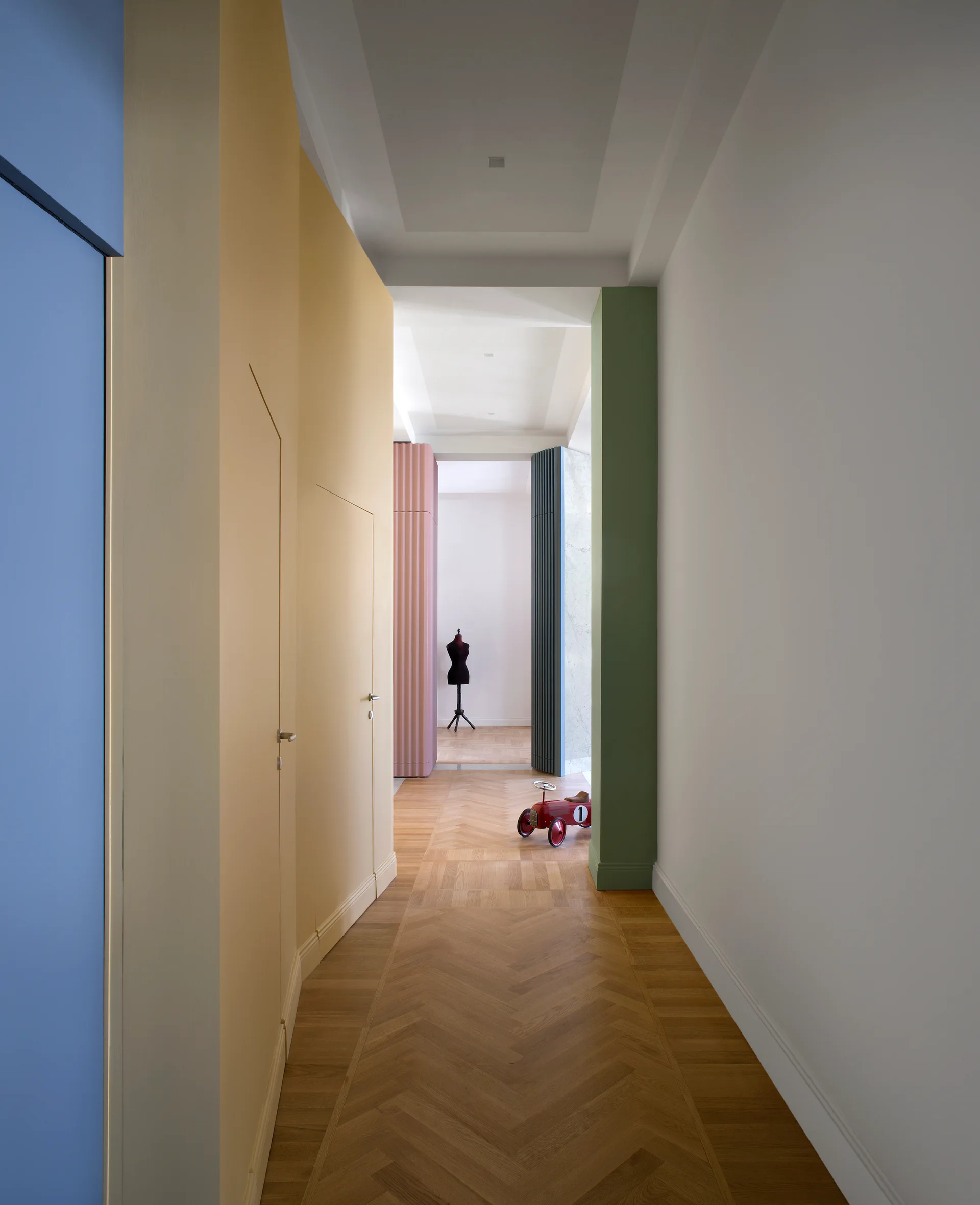 A long hallway featuring herringbone wood floors, a white ceiling, and matte walls color-blocked in blue, yellow, and green with flush-integrated doors.