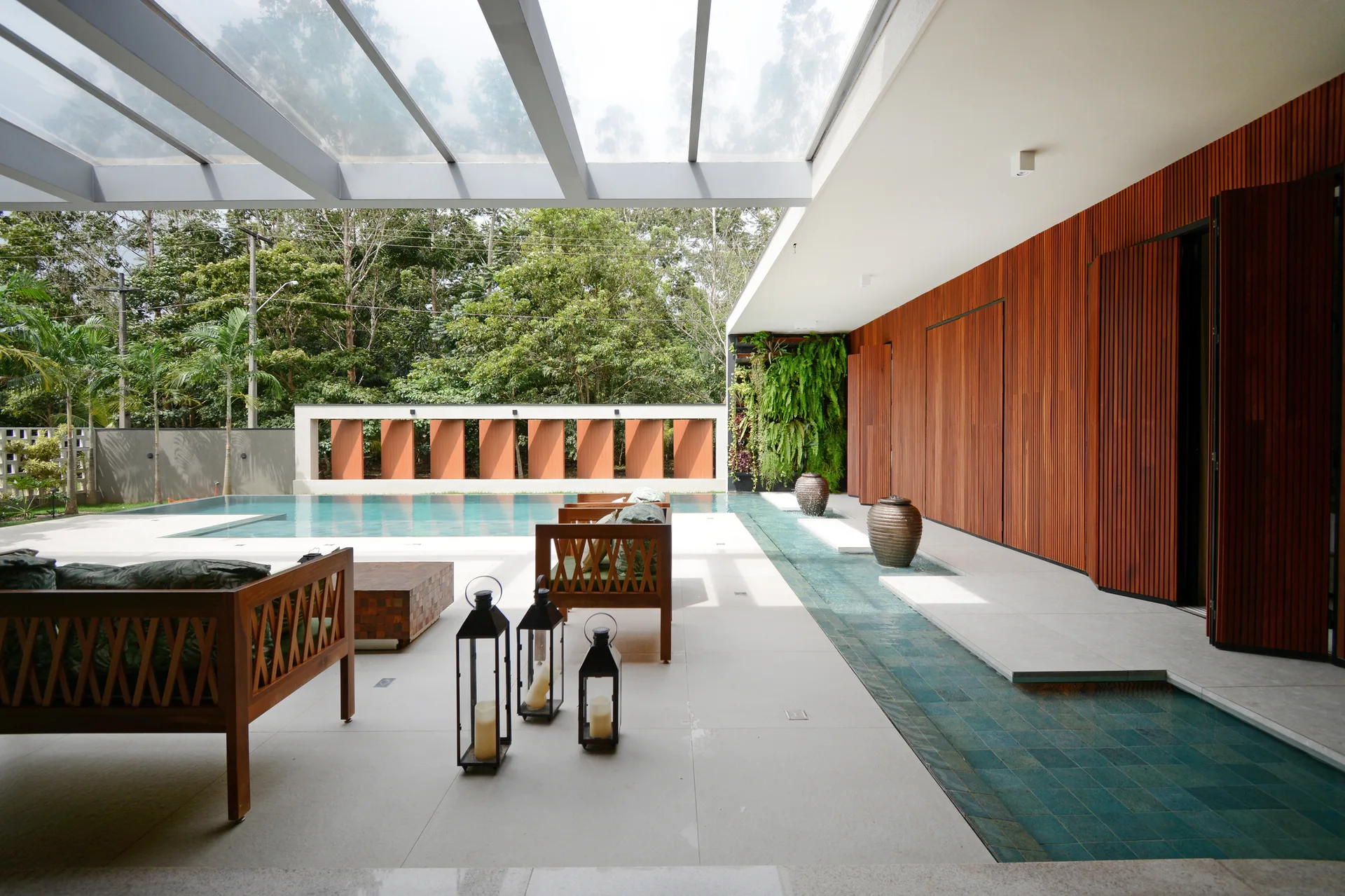 A modern covered patio features a glass canopy roof, wooden slat wall paneling, a large swimming pool, and wood frame sofas.