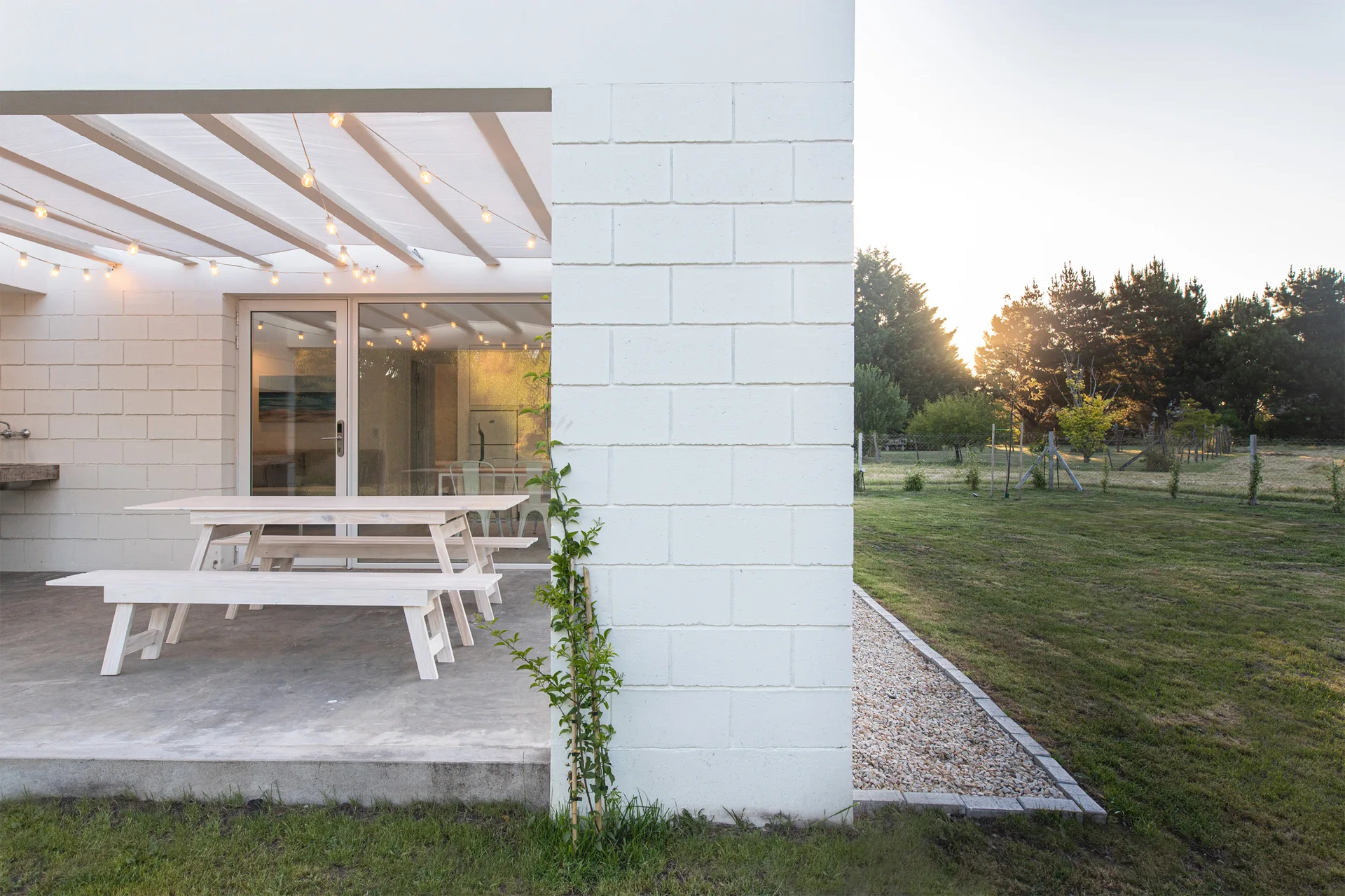 A covered concrete patio features a light wood picnic dining table and benches beneath a white pergola with string lights next to a white block wall.