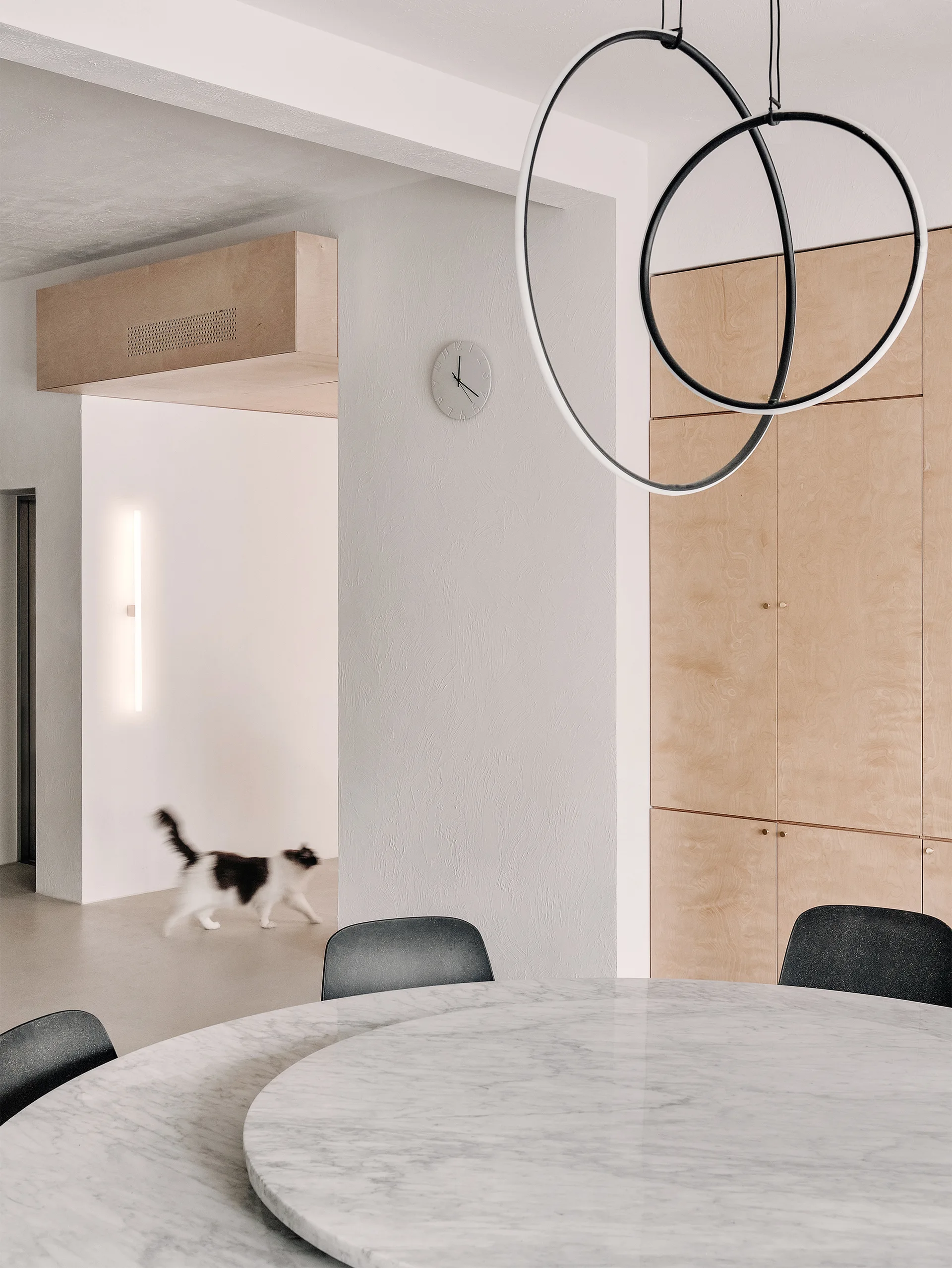 A minimalist dining room features a round marble table with black chairs, built-in plywood cabinets, and a black ring pendant light.