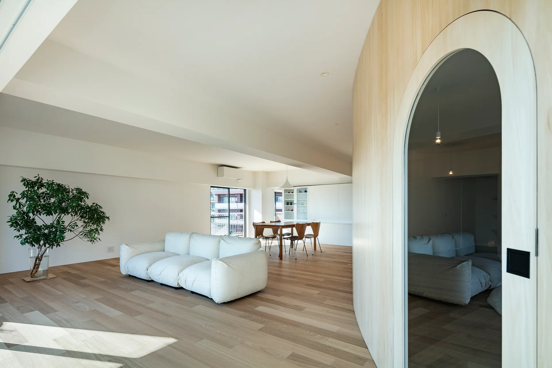 A minimalist living room with a low white sofa, light oak flooring, and a large curved wood partition wall with an arched glass door.