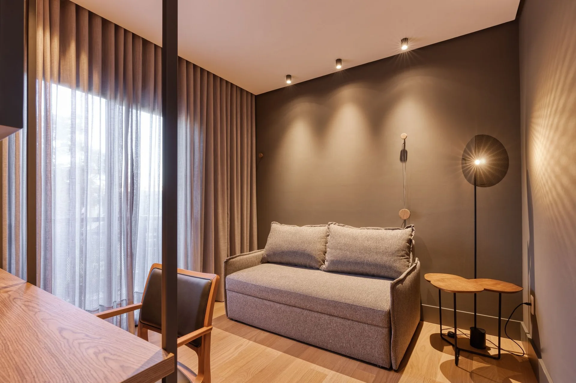 A gray sofa bed stands against a dark gray wall illuminated by warm ceiling spotlights, beside a wooden desk and a sheer curtained window.