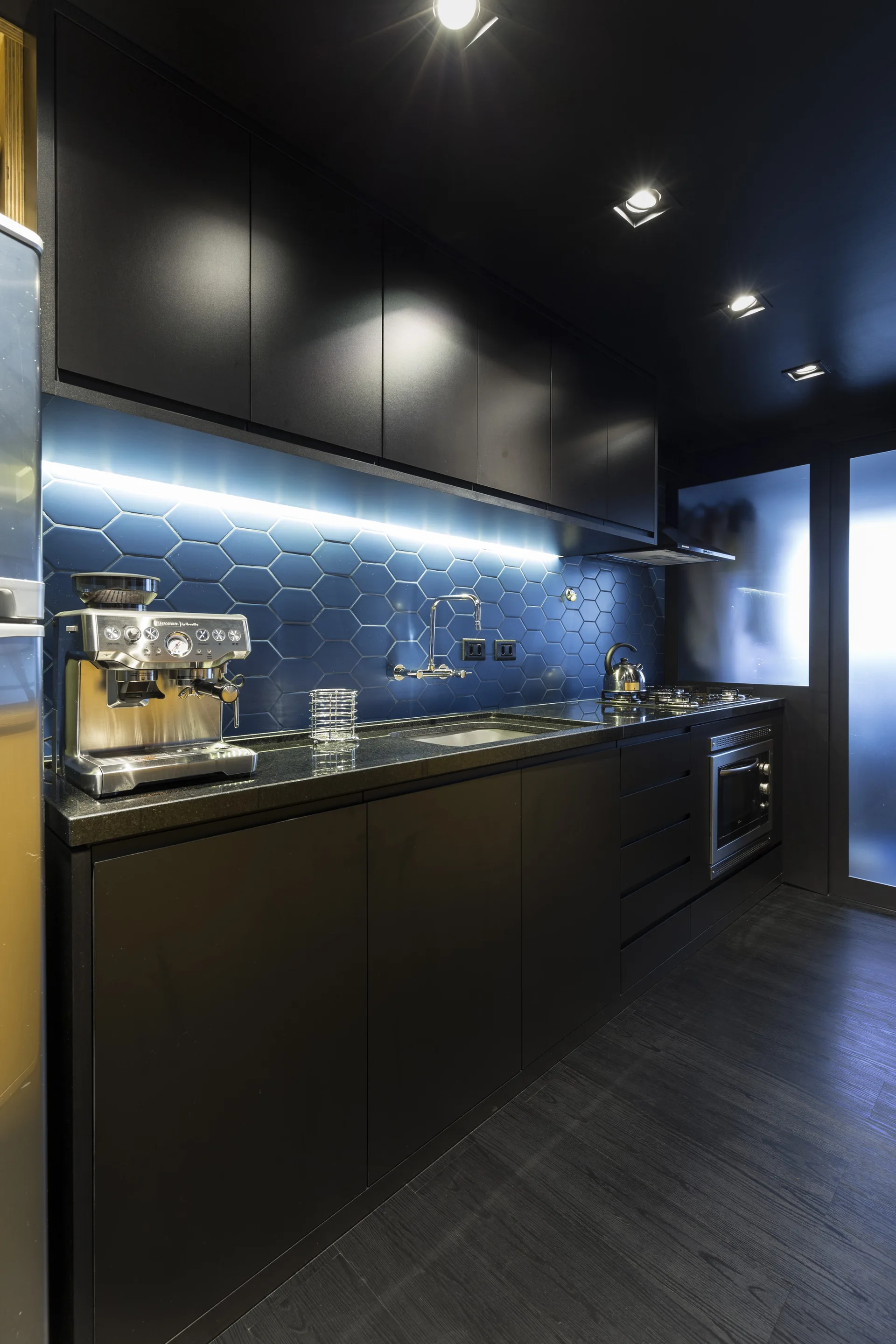 Matte black kitchen cabinets and countertops frame a glossy blue hexagonal tile backsplash illuminated by an LED strip.