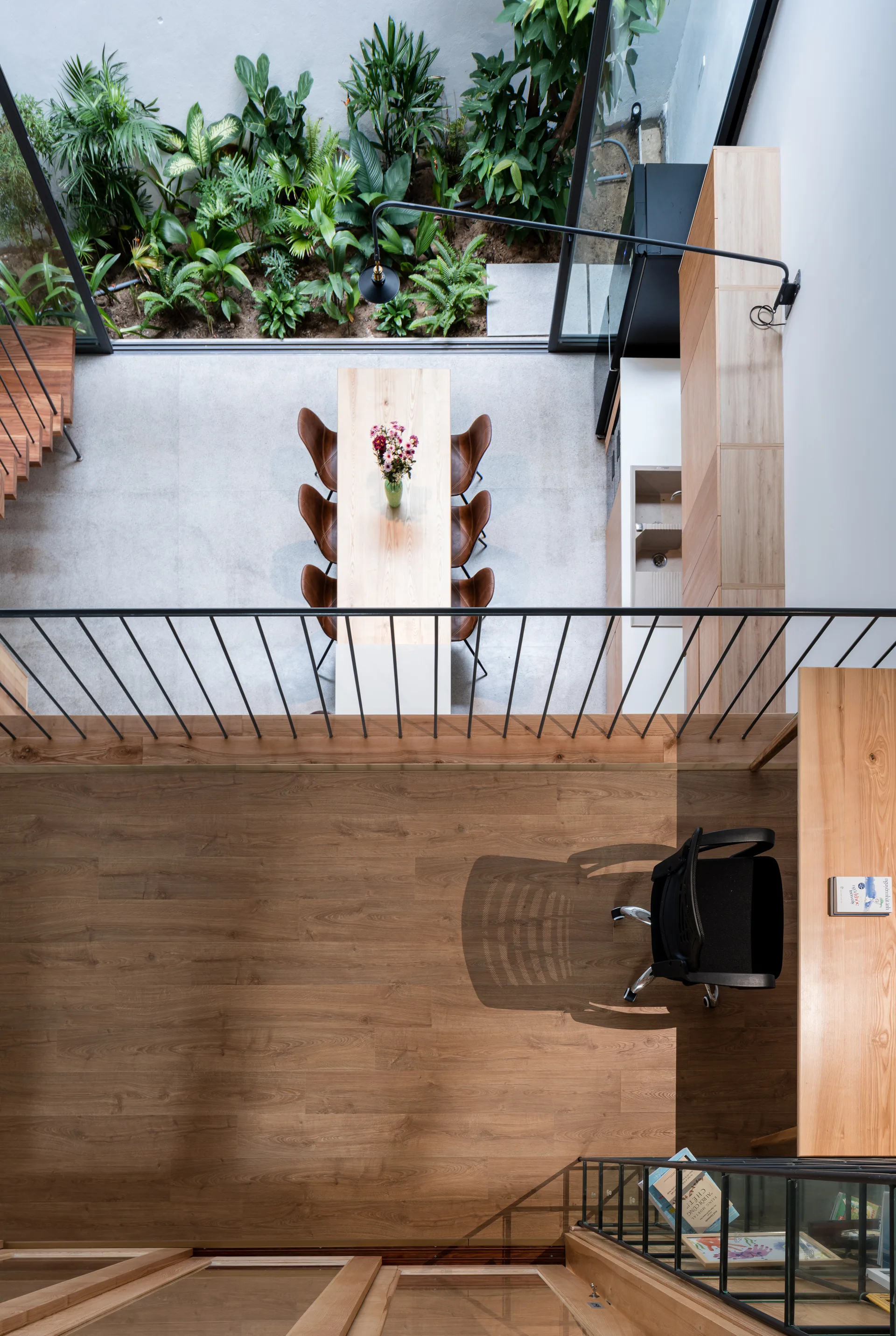 An overhead view from a wooden mezzanine workspace looking down at a ground-floor dining table and a glass-walled indoor courtyard garden.