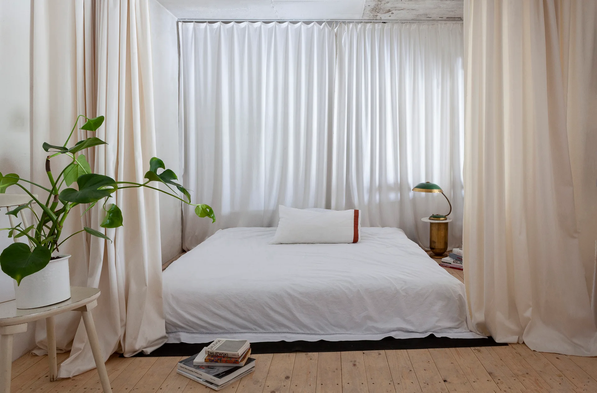 A low-profile bed with white linens is nestled behind off-white curtains on light wood floors, accented by a brass lamp and a potted monstera plant.