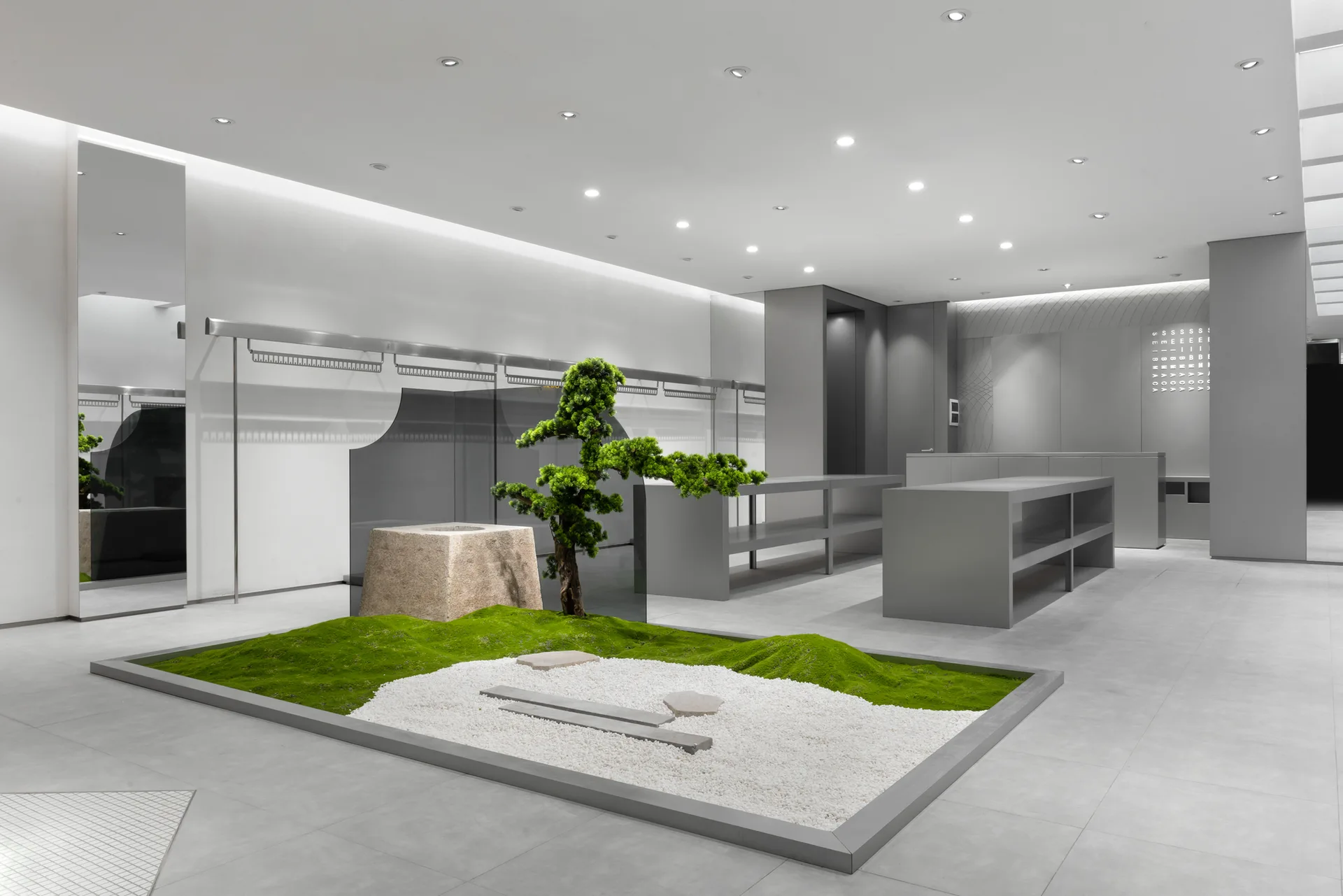 A minimalist gray retail space featuring a central indoor zen garden with green moss, white gravel, and a small shaped tree next to gray display tables.