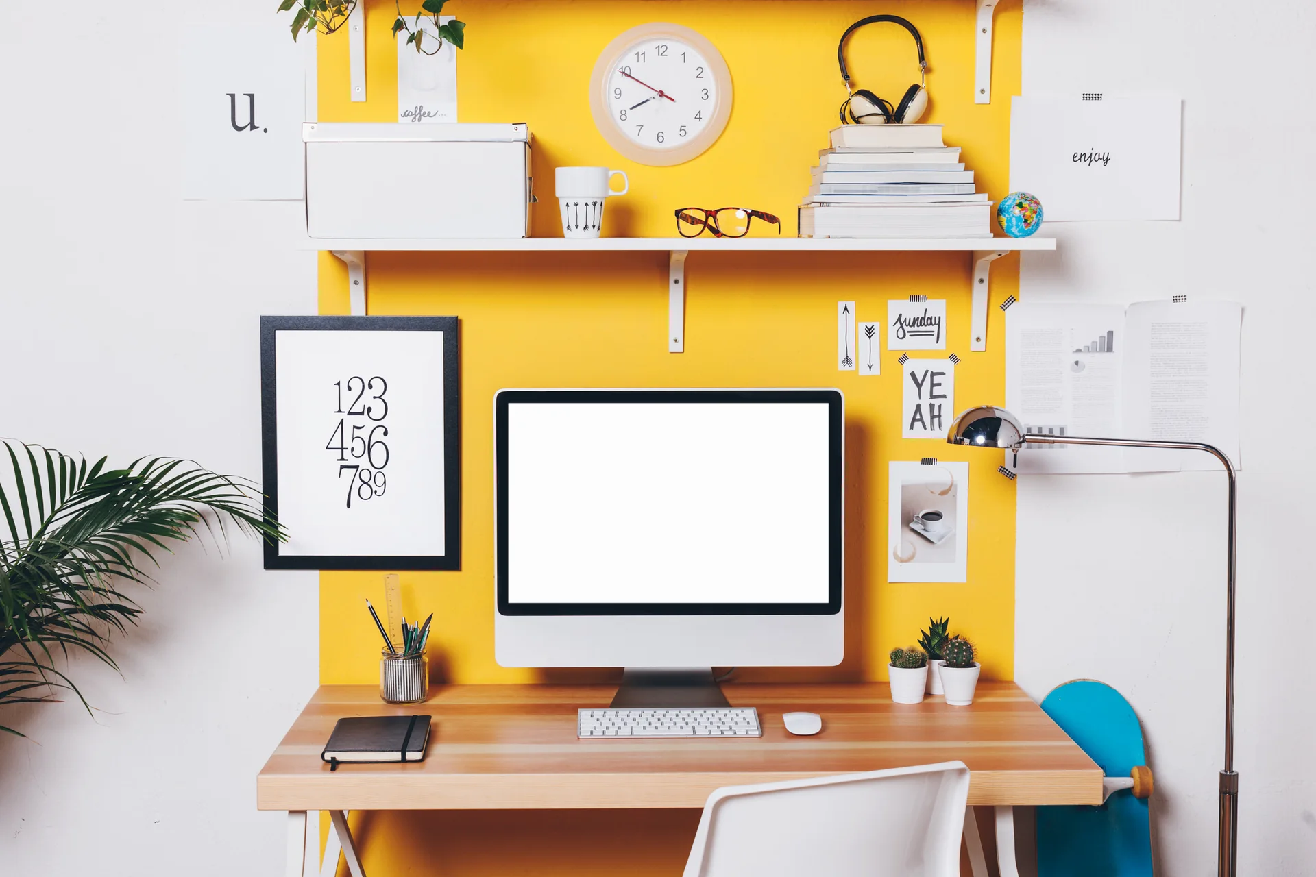 A wooden desk with a computer sits in front of a bright yellow vertical accent stripe on a white wall, topped by a white display shelf.