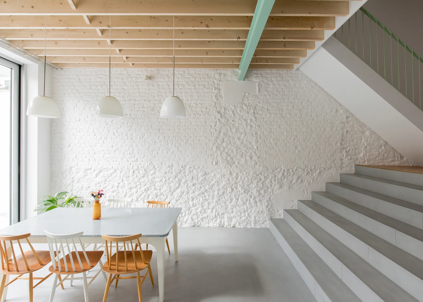 A modern dining room features a light gray table with wooden chairs, three white pendant lights, a textured brick wall, and an exposed wood ceiling.