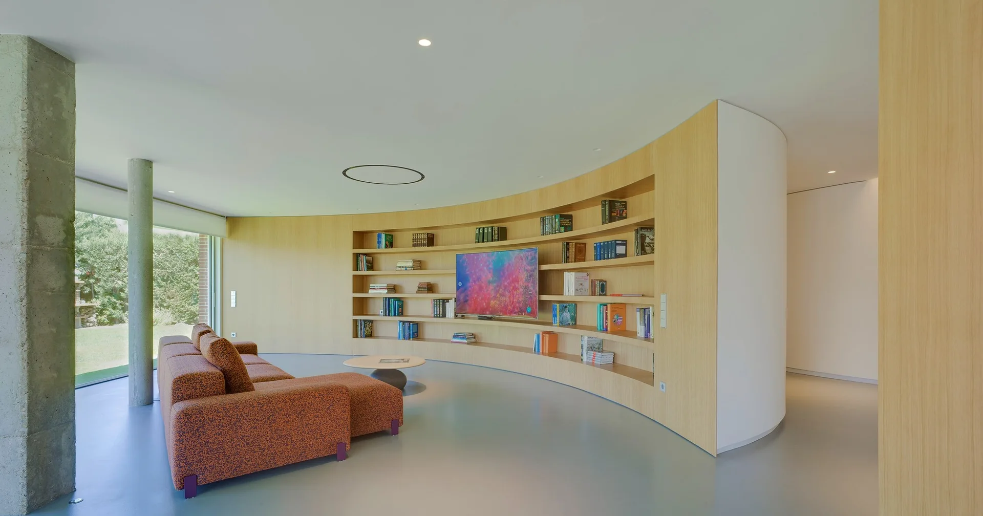 A modern living room featuring a curved light wood built-in bookshelf wall, an orange textured sofa, a round coffee table, and a concrete column.