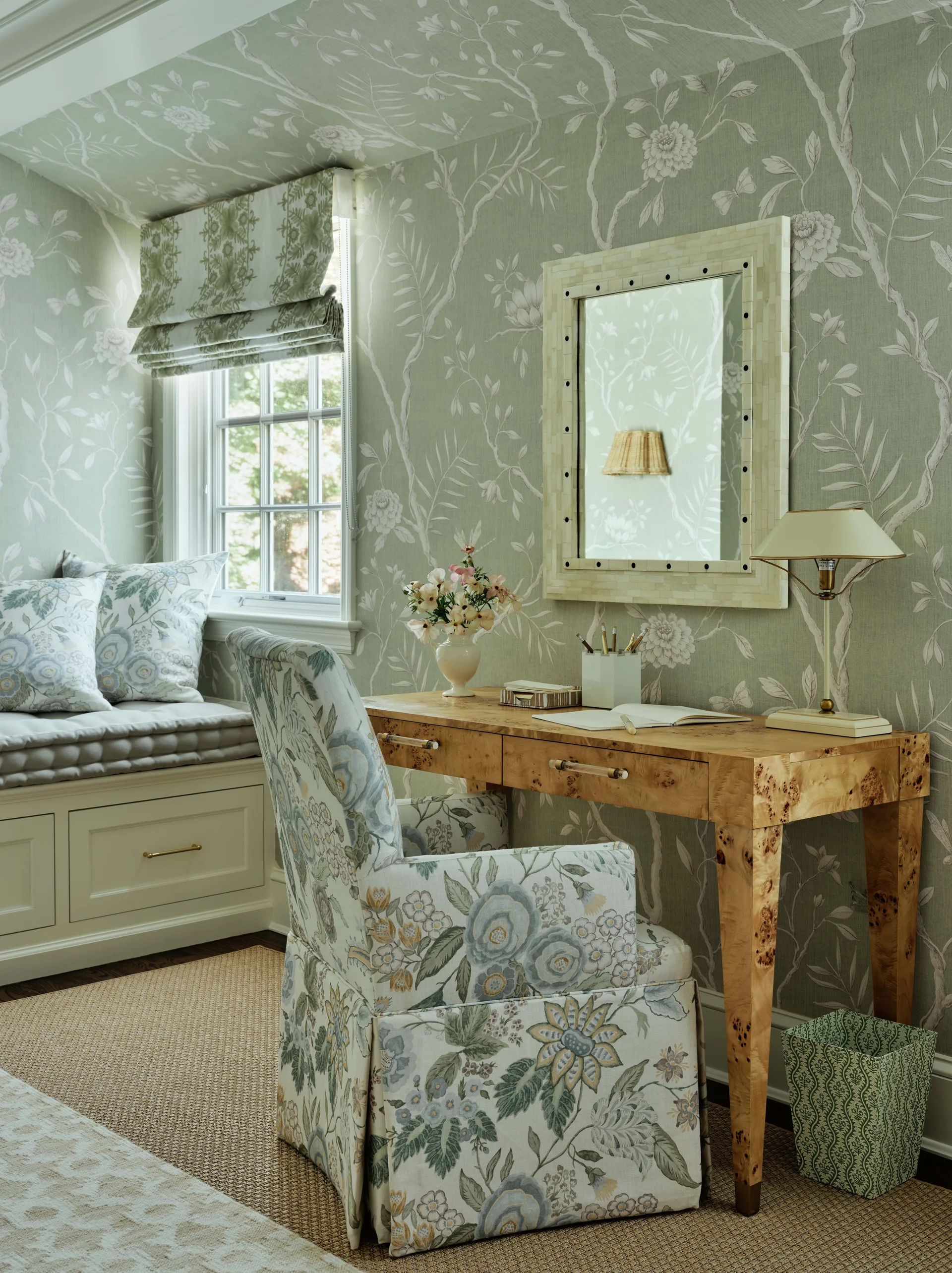 A home office features sage green floral wallpaper, a burl wood desk with an upholstered armchair, and a built-in window seat under a roman shade.