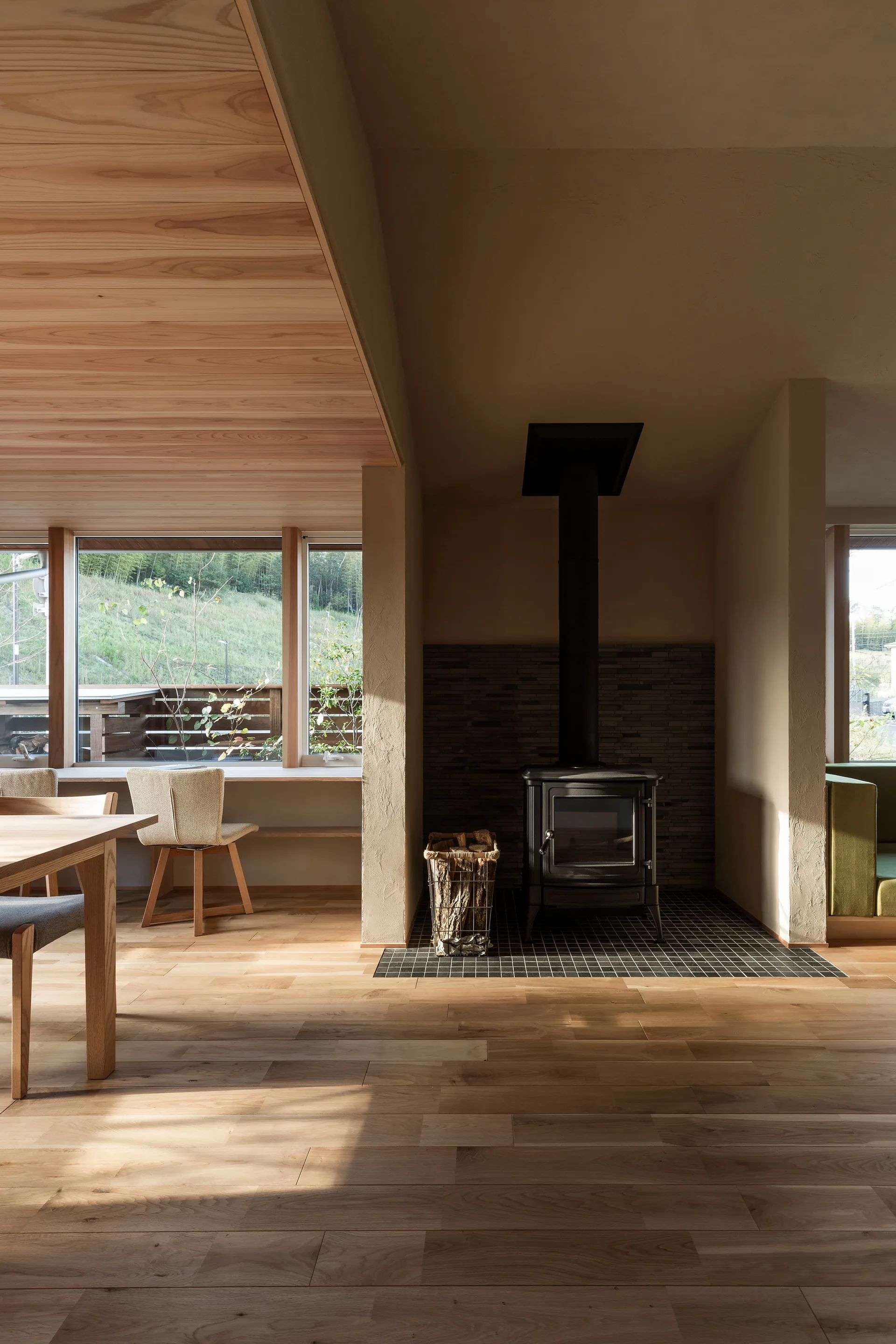 A black wood stove sits on a tiled hearth in a sunlit room featuring light wood floors, plaster walls, and a timber plank ceiling.