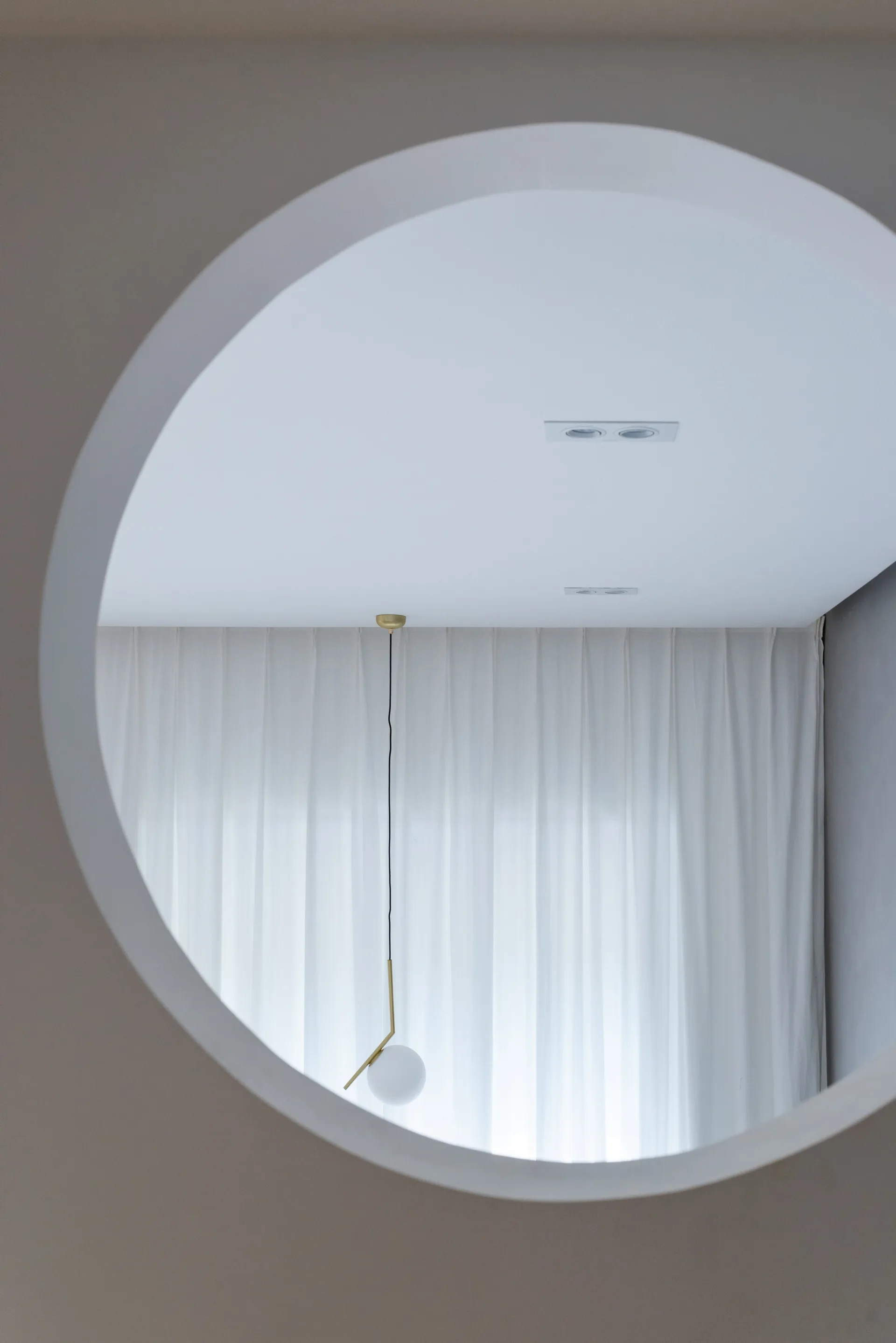 A view through a circular wall opening reveals a minimalist globe pendant light hanging in front of sheer white drapery.