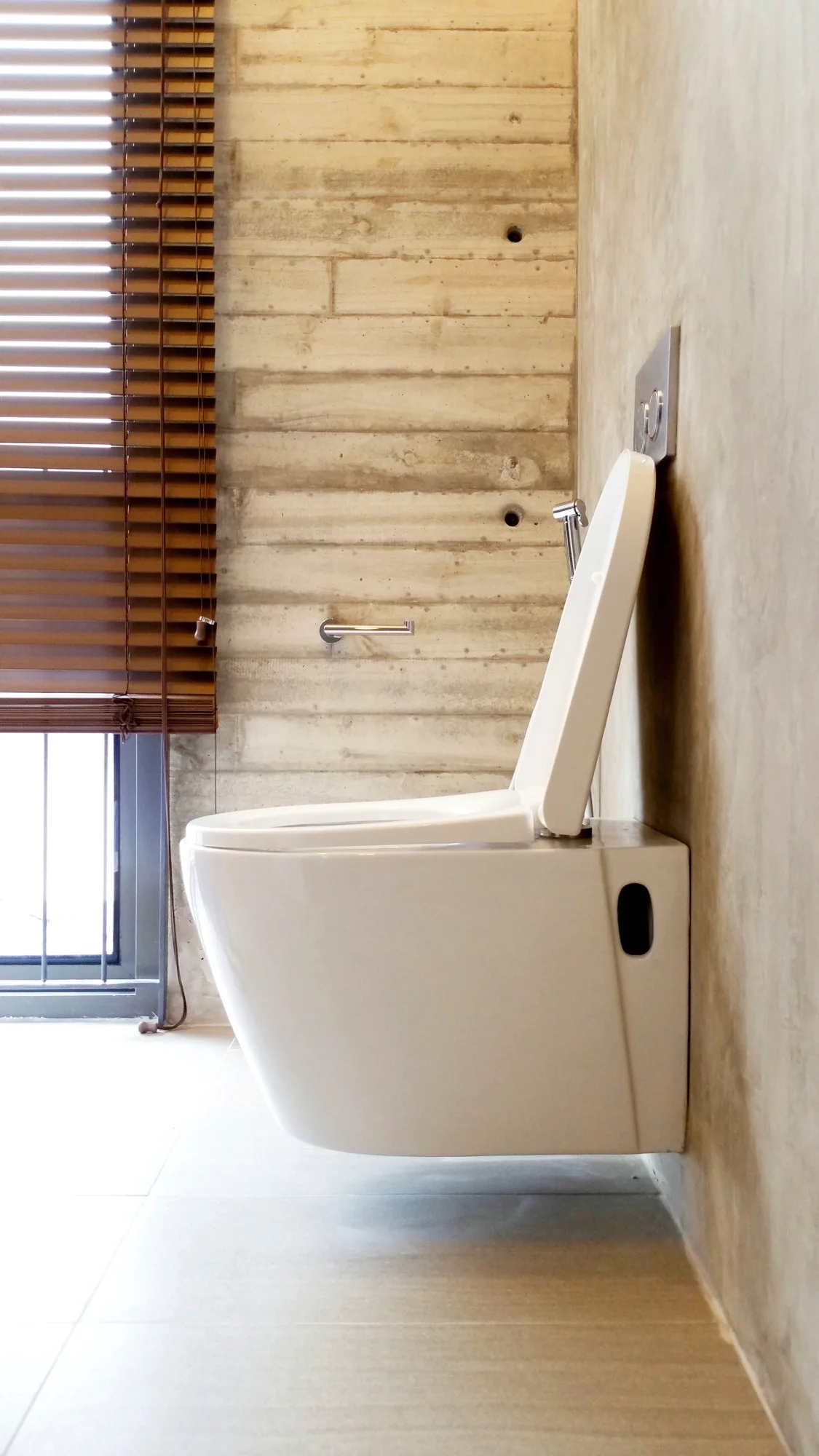 A white wall-mounted toilet is fixed to a board-formed concrete wall next to a window with brown wooden blinds.