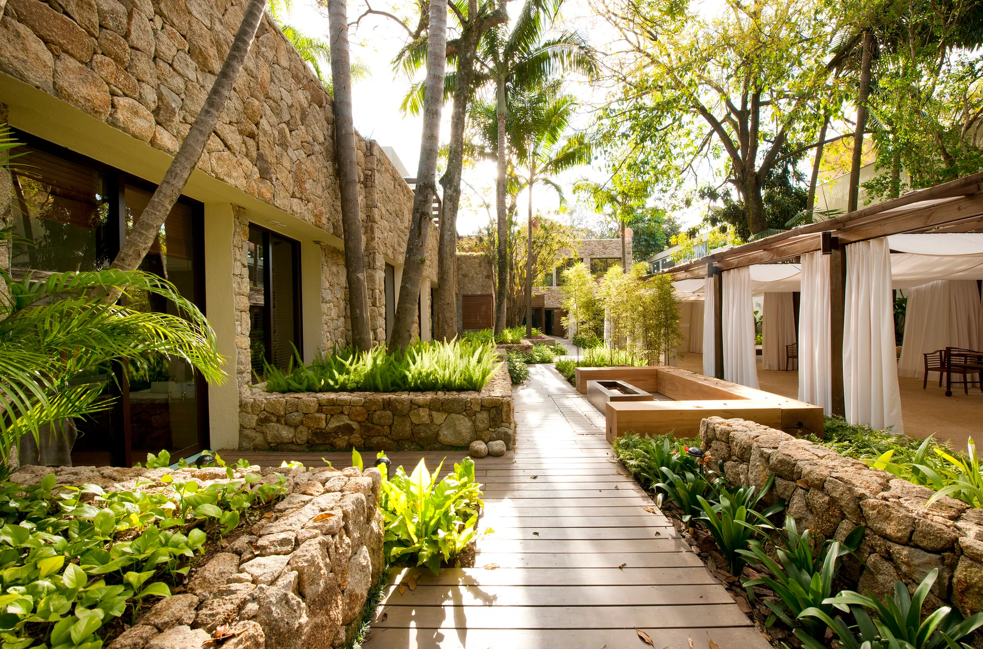 A sunlit wooden walkway runs through a tropical courtyard lined with rustic stacked stone planter beds and a white-curtained pergola.
