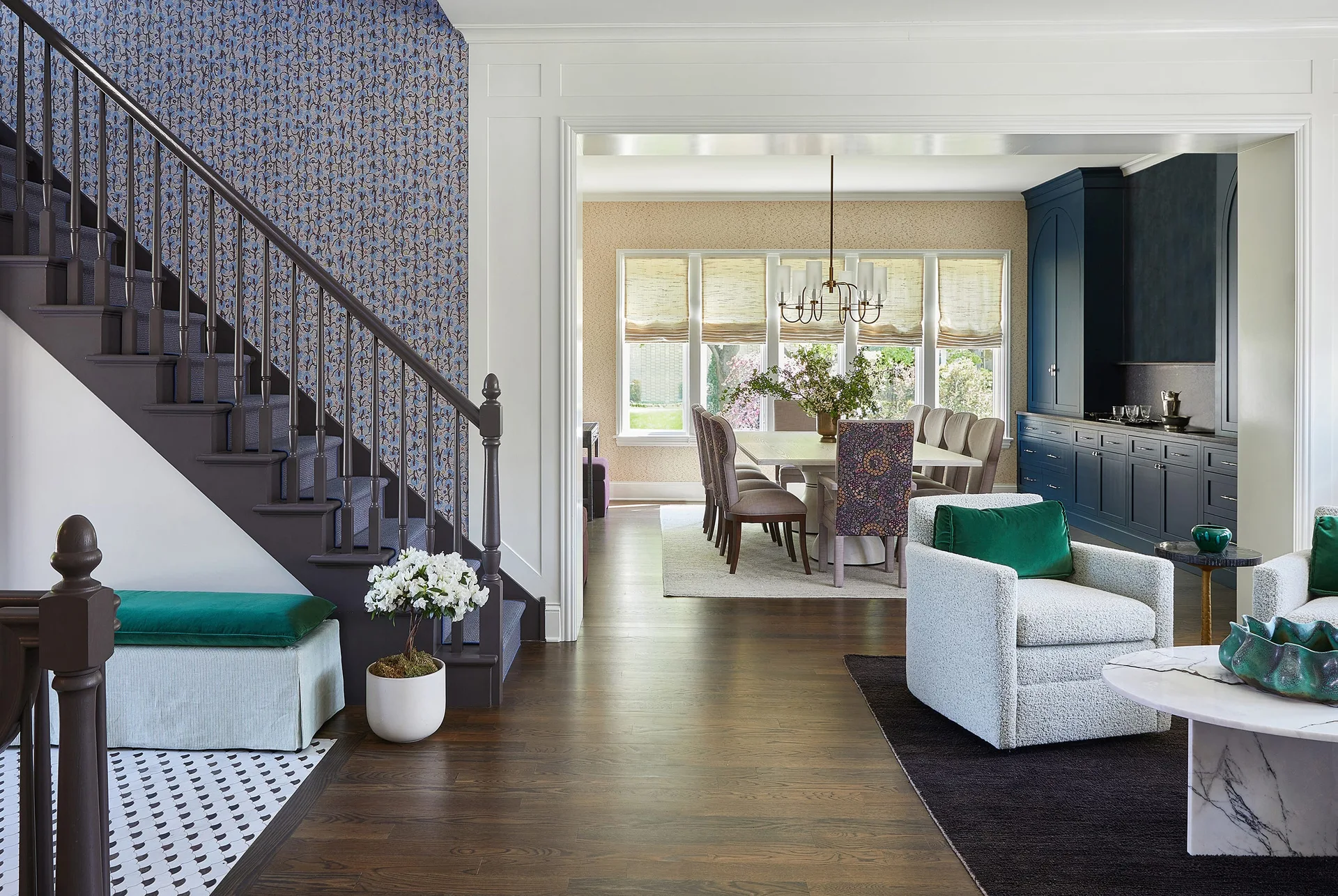 An entryway with a dark wood staircase and patterned blue wallpaper transitions into a bright dining room with dark blue cabinetry.