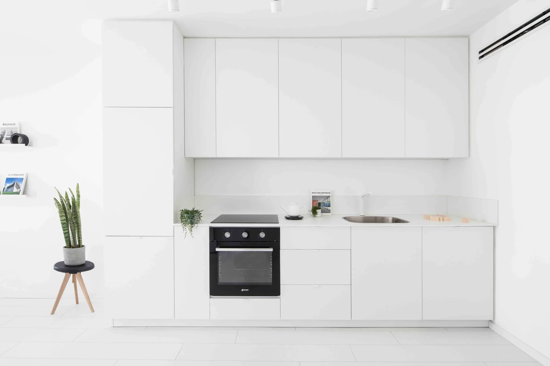 A minimalist white kitchen featuring flat-front handleless cabinets, a built-in black oven, a small stainless steel sink, and a potted plant.