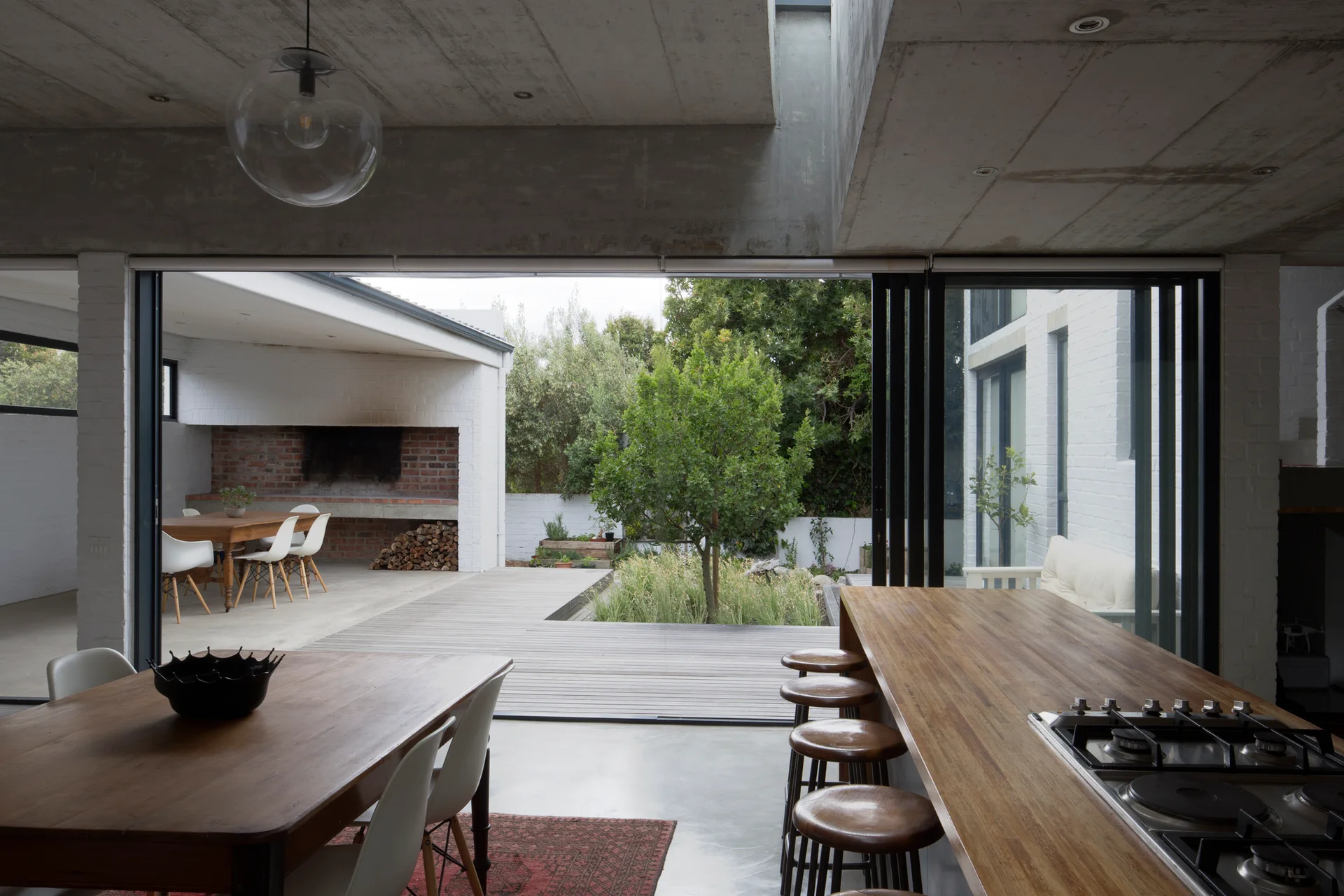 A modern dining area features an exposed concrete ceiling, a wooden breakfast bar with stools, and sliding glass doors that open to a wood deck.