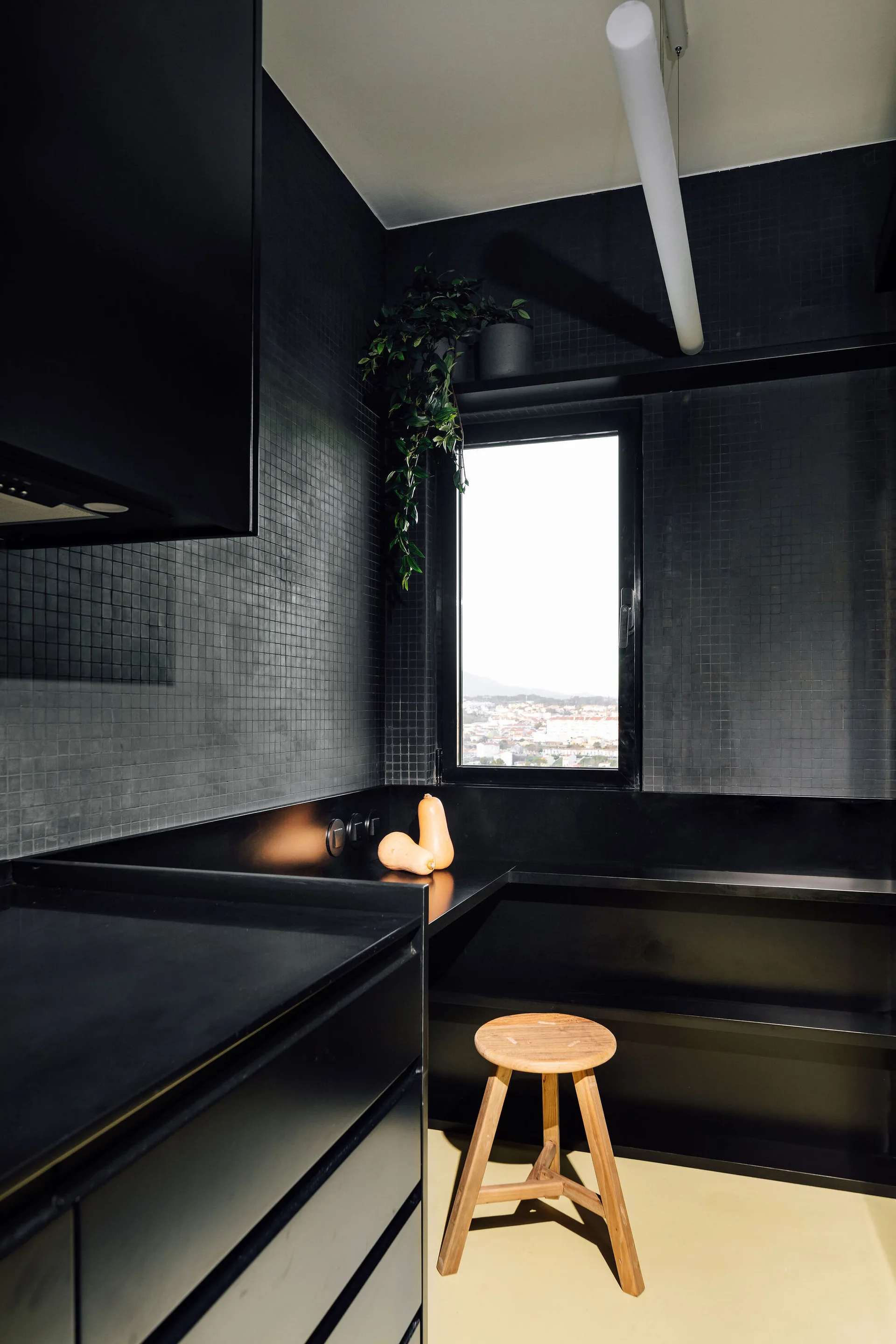 A modern kitchen features matte black tiled walls, sleek black countertops, and a three-legged wooden stool on a light-colored floor.