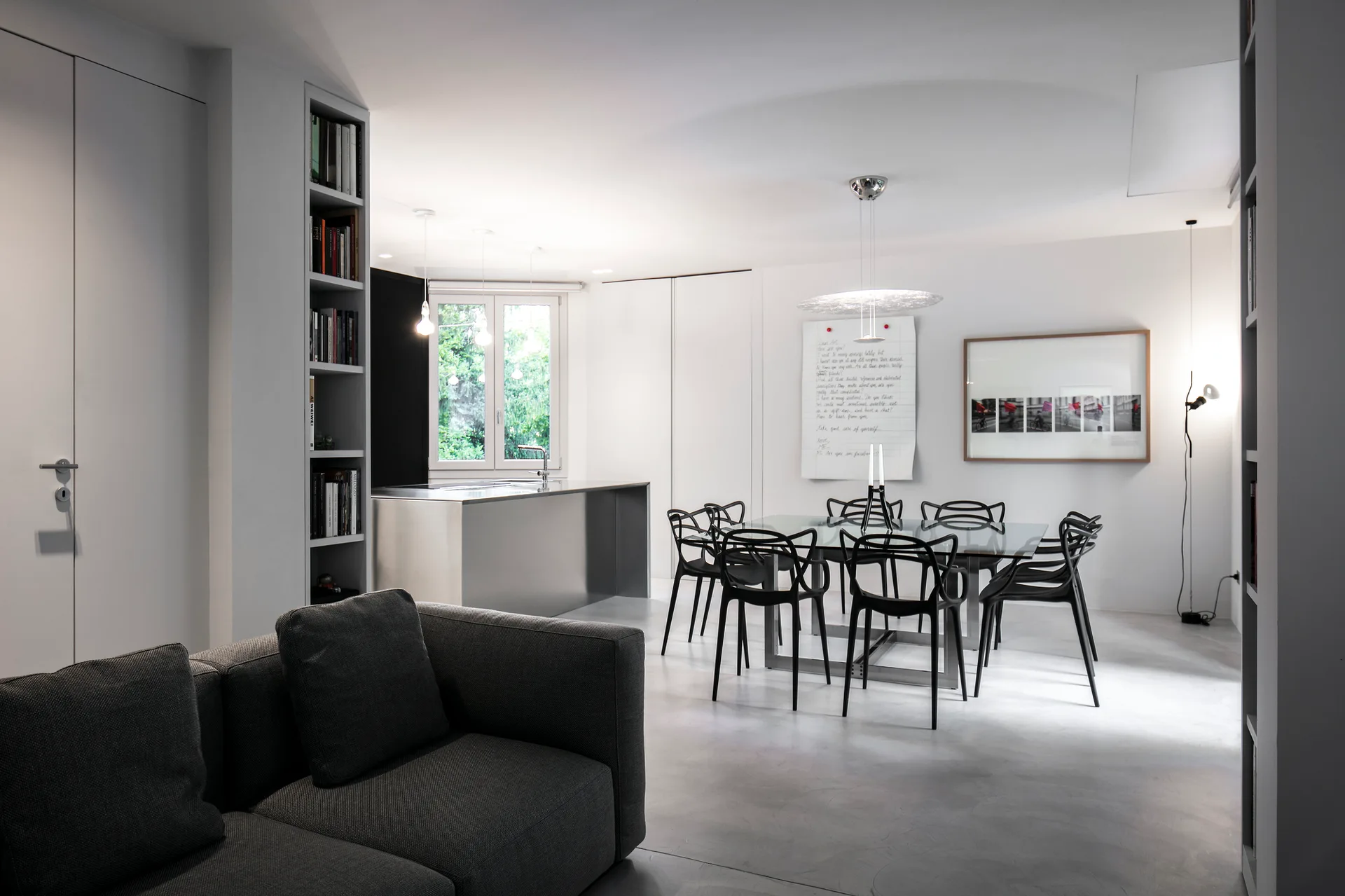 A minimalist dining room features a glass-top table surrounded by black sculptural chairs, situated near a metallic kitchen island on a grey floor.
