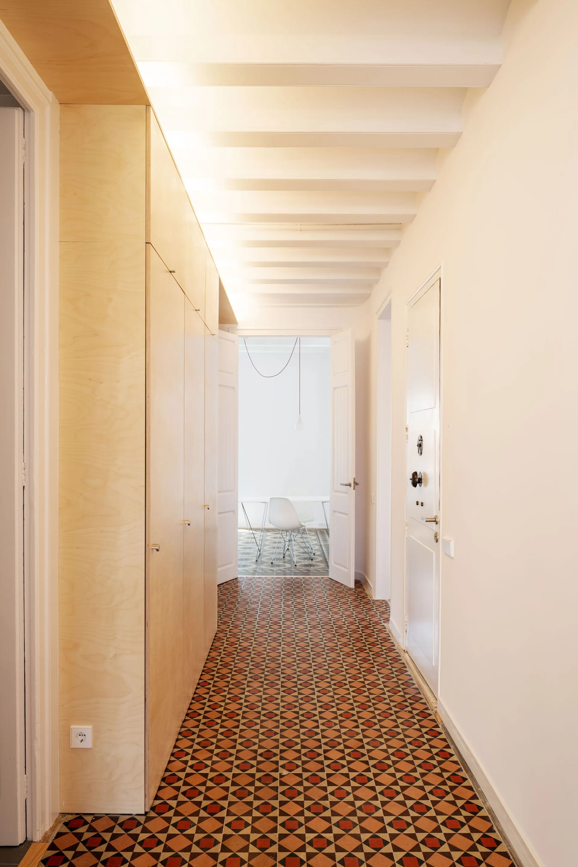 A bright residential hallway featuring floor-to-ceiling light wood cabinetry, white painted ceiling beams, and a geometric patterned tile floor.