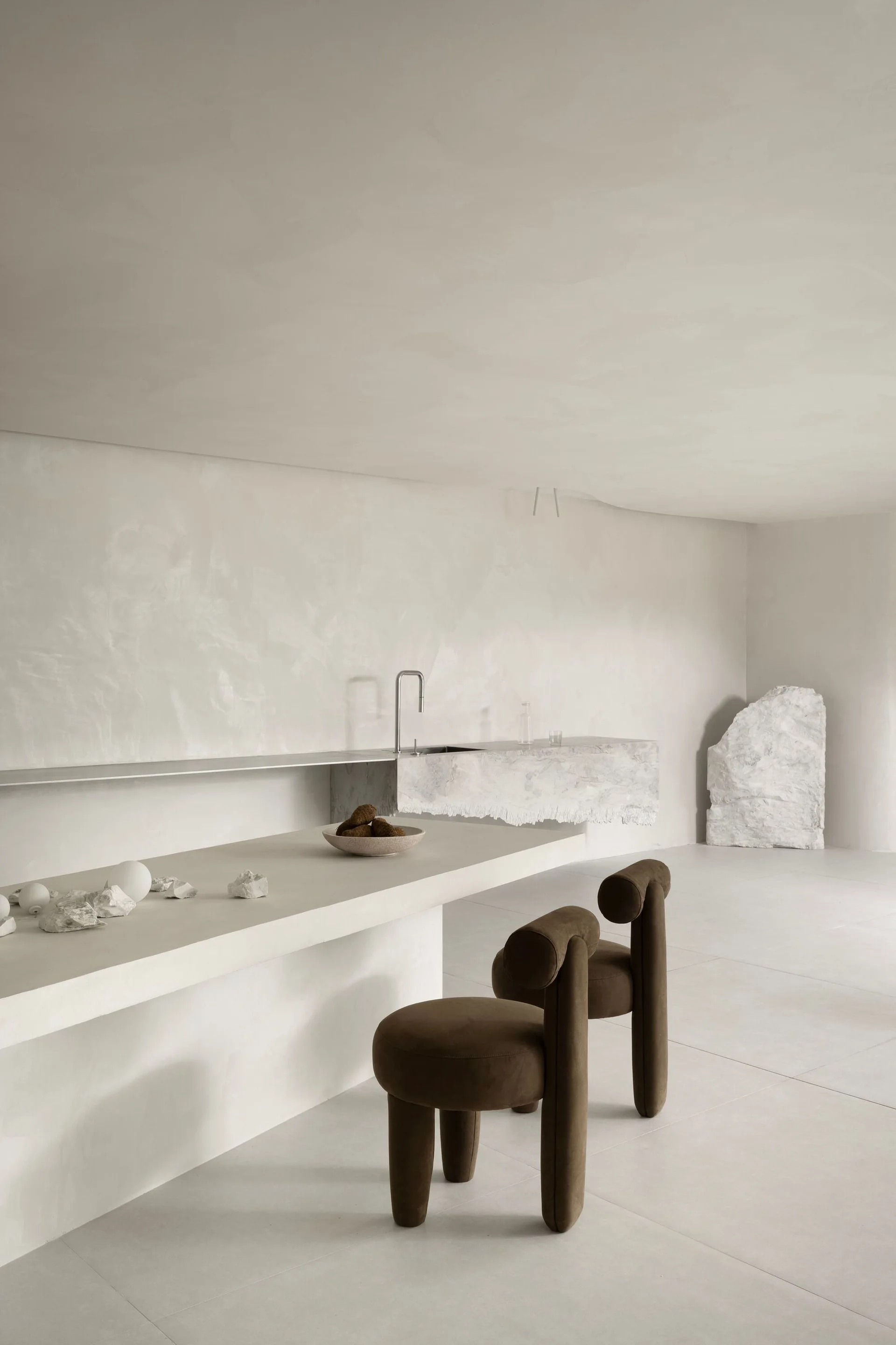 A minimalist off-white kitchen features a long plastered island, two brown sculptural stools, a cantilevered stone sink counter, and textured walls.