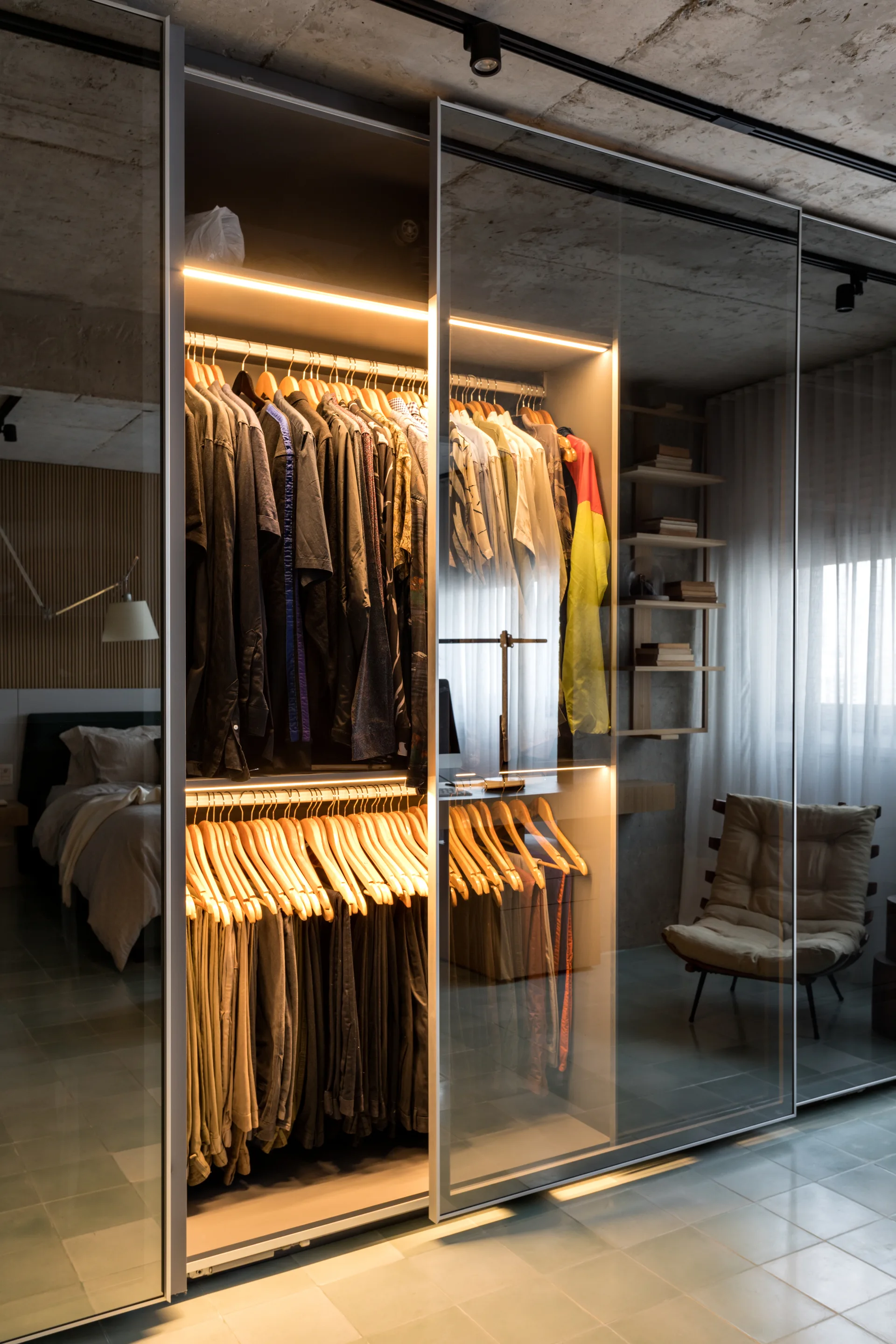 A bedroom features a large wardrobe with glass sliding doors, lit internally by warm LED strips, against an exposed concrete ceiling.