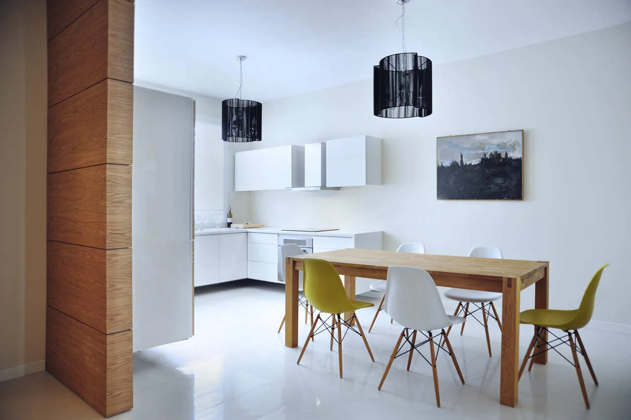 A wooden dining table with white and mustard yellow chairs sits in a bright white open kitchen featuring two black pendant lights and a wood partition.