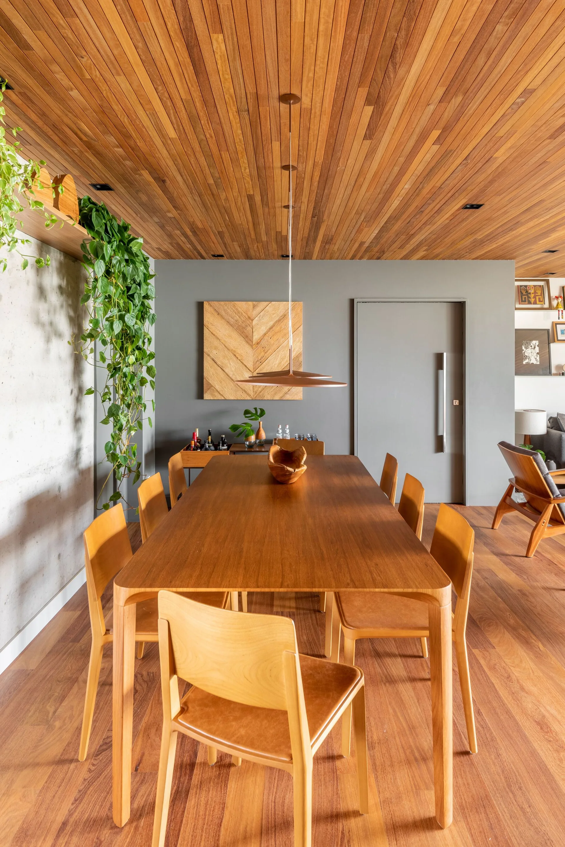 A modern dining room features a long wooden table with matching chairs under a wood slat ceiling next to a grey accent wall and concrete wall.